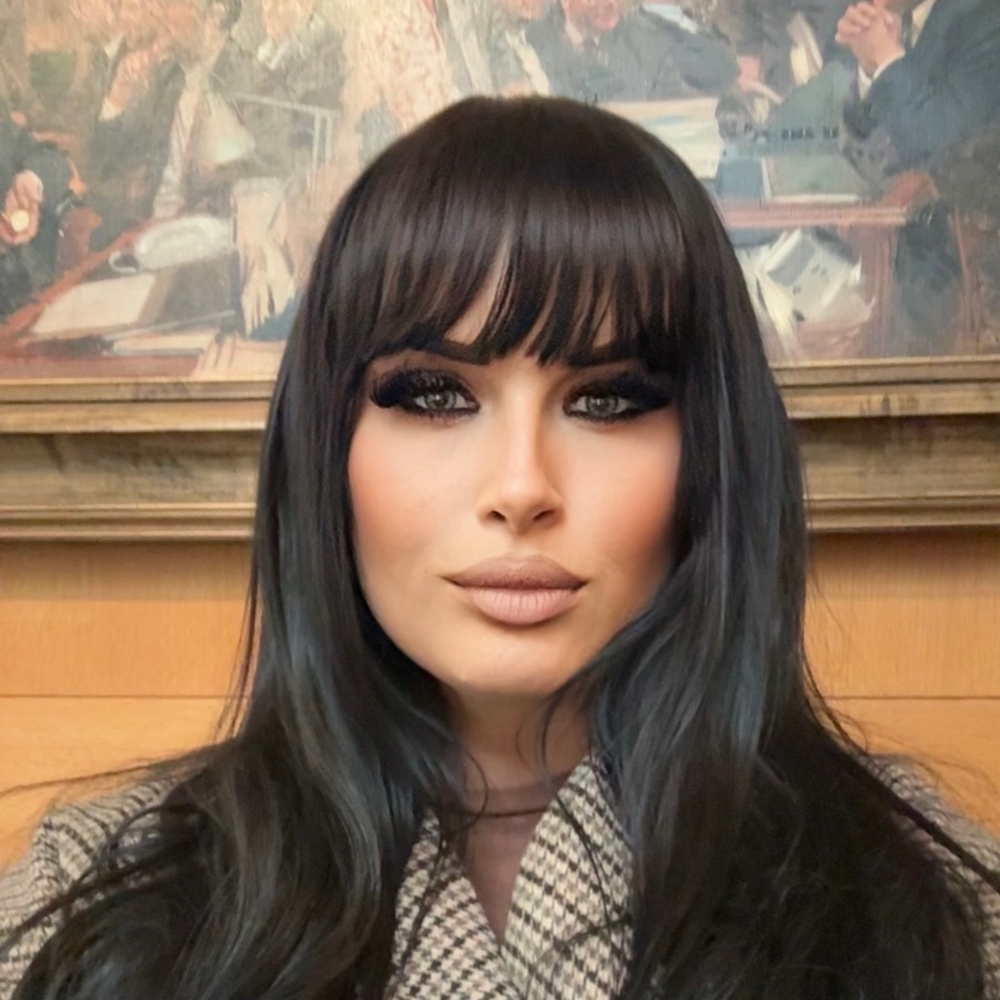Close-up of a woman with long dark hair and bangs, wearing makeup, sitting in front of a painting in a room with wood walls.
