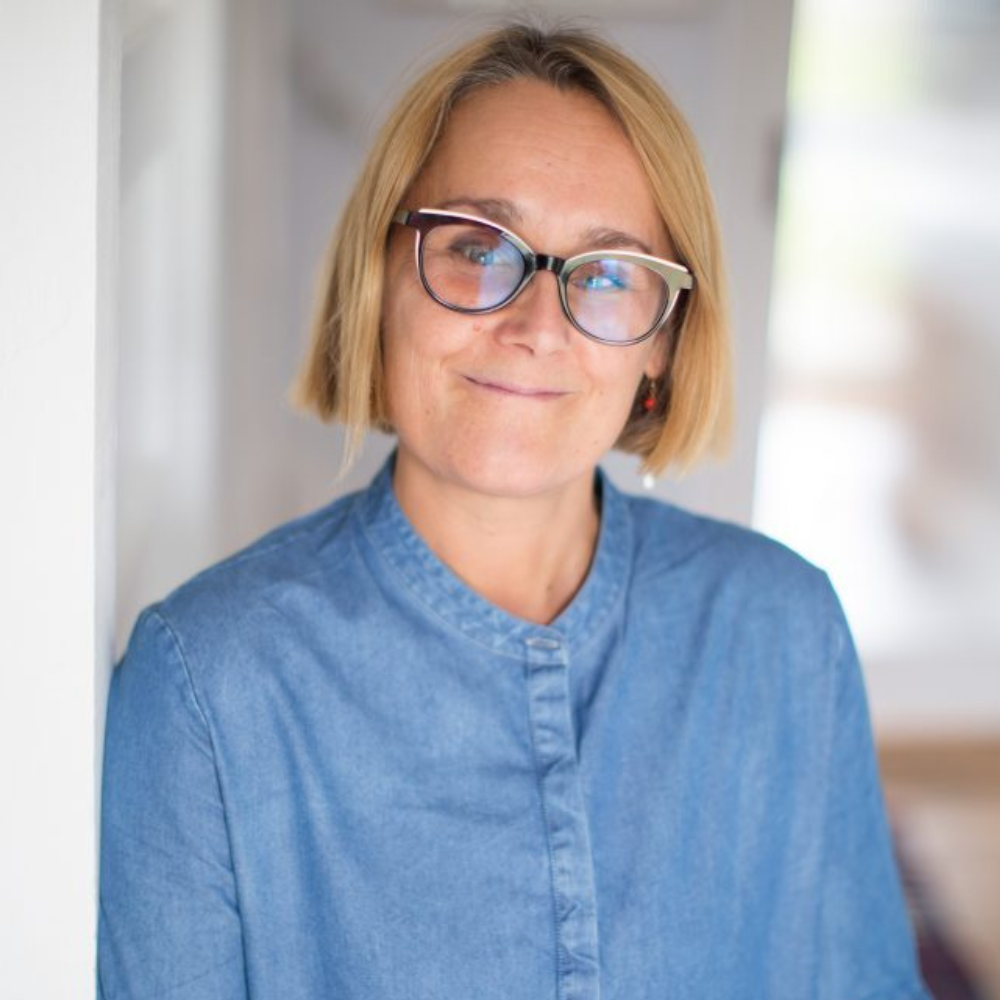 A woman with blonde hair, glasses, and a blue denim shirt smiling and leaning against a wall in a well-lit indoor space.
