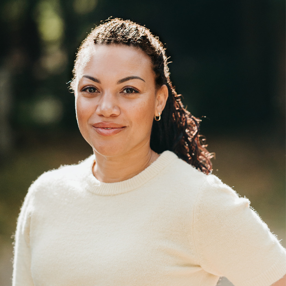 A woman with curly hair, wearing a cream sweater, standing outdoors in natural light.