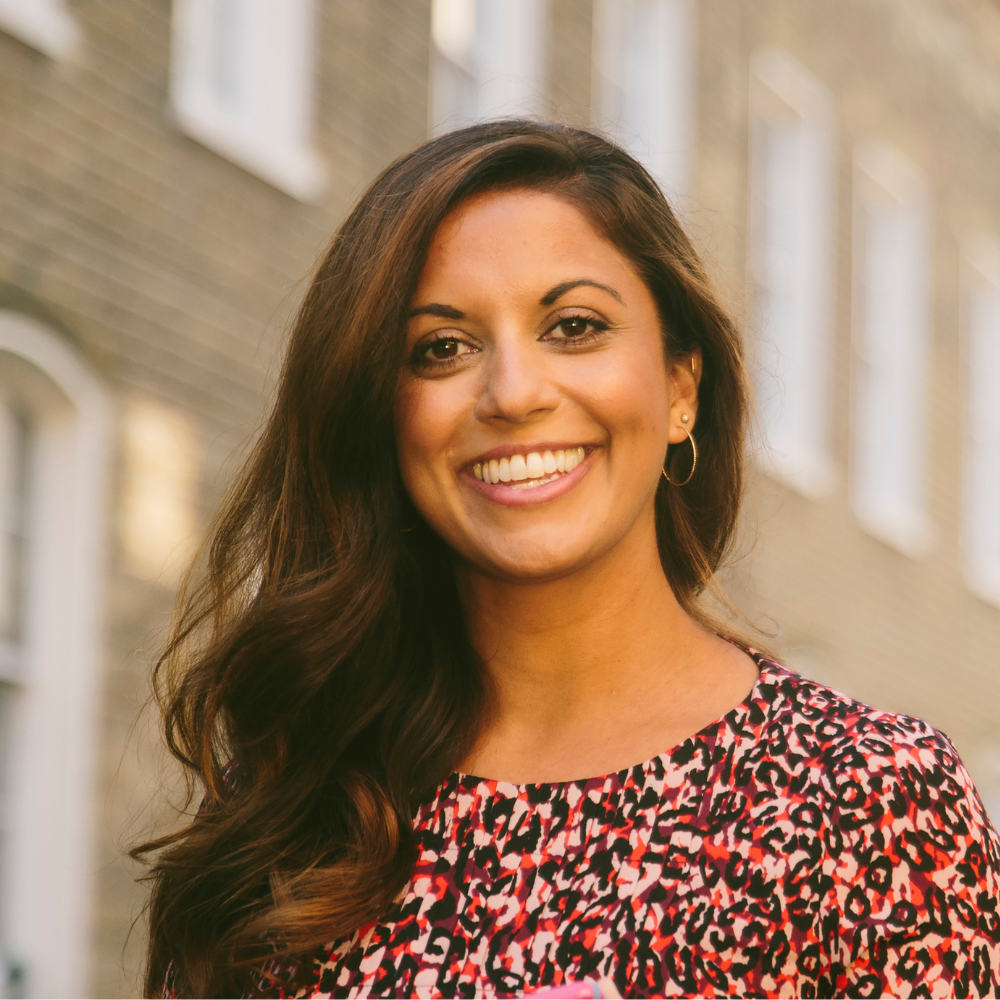 A woman with long dark hair smiling outdoors in front of brick buildings, wearing a colorful floral top and hoop earrings.