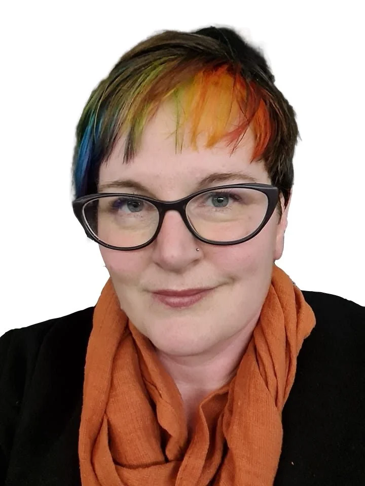 Person with short hair and rainbow-colored bangs wearing glasses, a black top, and an orange scarf, smiling against a white background.