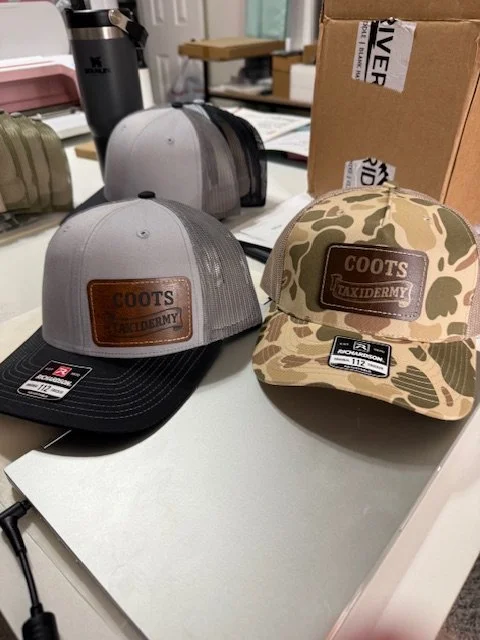 Two baseball caps on a desk, one with a gray and black camouflage pattern, and the other with a gray front and camouflage mesh back, both with leather patches that say 'COOTS TACKLDERMY'. In the background, there are boxes, papers, and a coffee thermos.