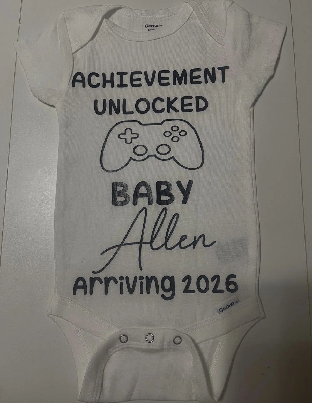 White baby onesie with black text that reads "Achievement Unlocked Baby Allen arriving 2026" and a graphic of a video game controller.