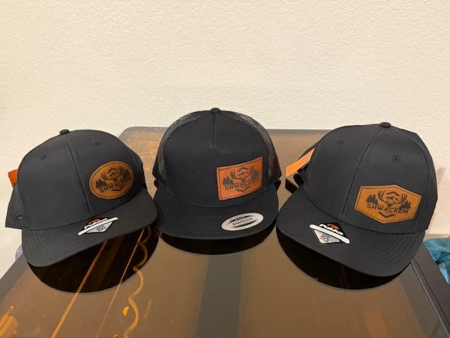 Three black snapback hats with a tan logo patch reading 'SHWACKER' and showing a mountain landscape, placed on a reflective surface against a beige wall.