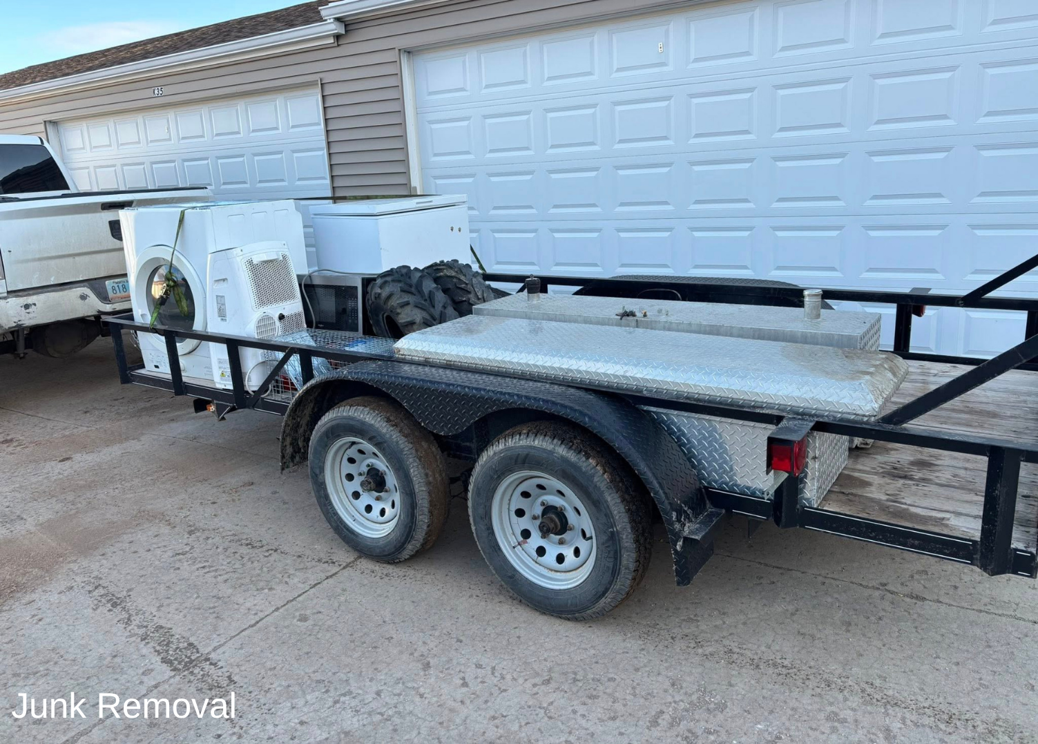 Trailer loaded with household appliances, tires, and metal boxes parked in front of a garage.