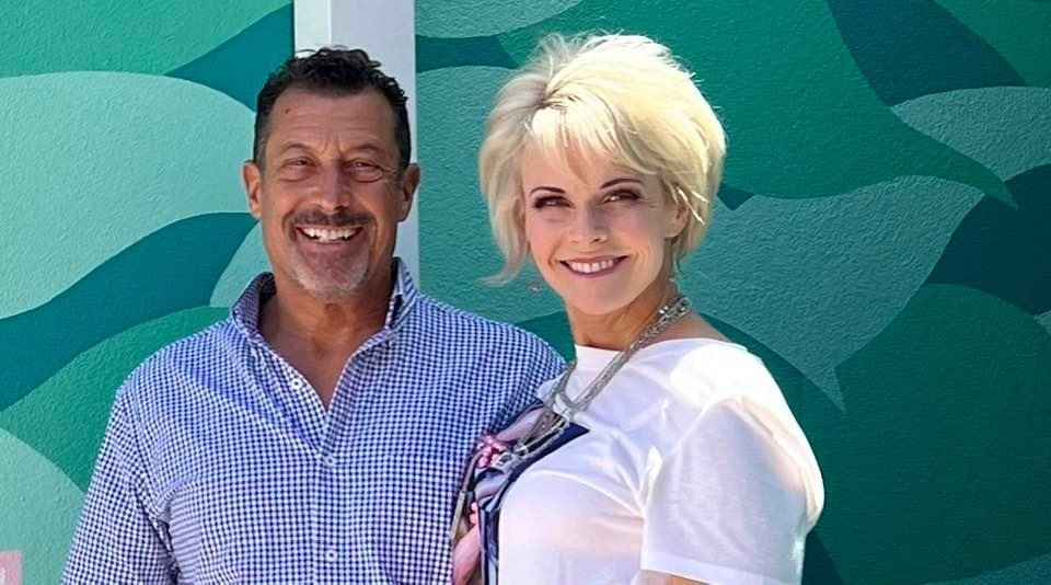 A smiling middle-aged man with dark hair and a beard wearing a blue checked shirt standing next to a smiling woman with short blonde hair in a white shirt, standing in front of a colorful mural with abstract green and blue shapes.