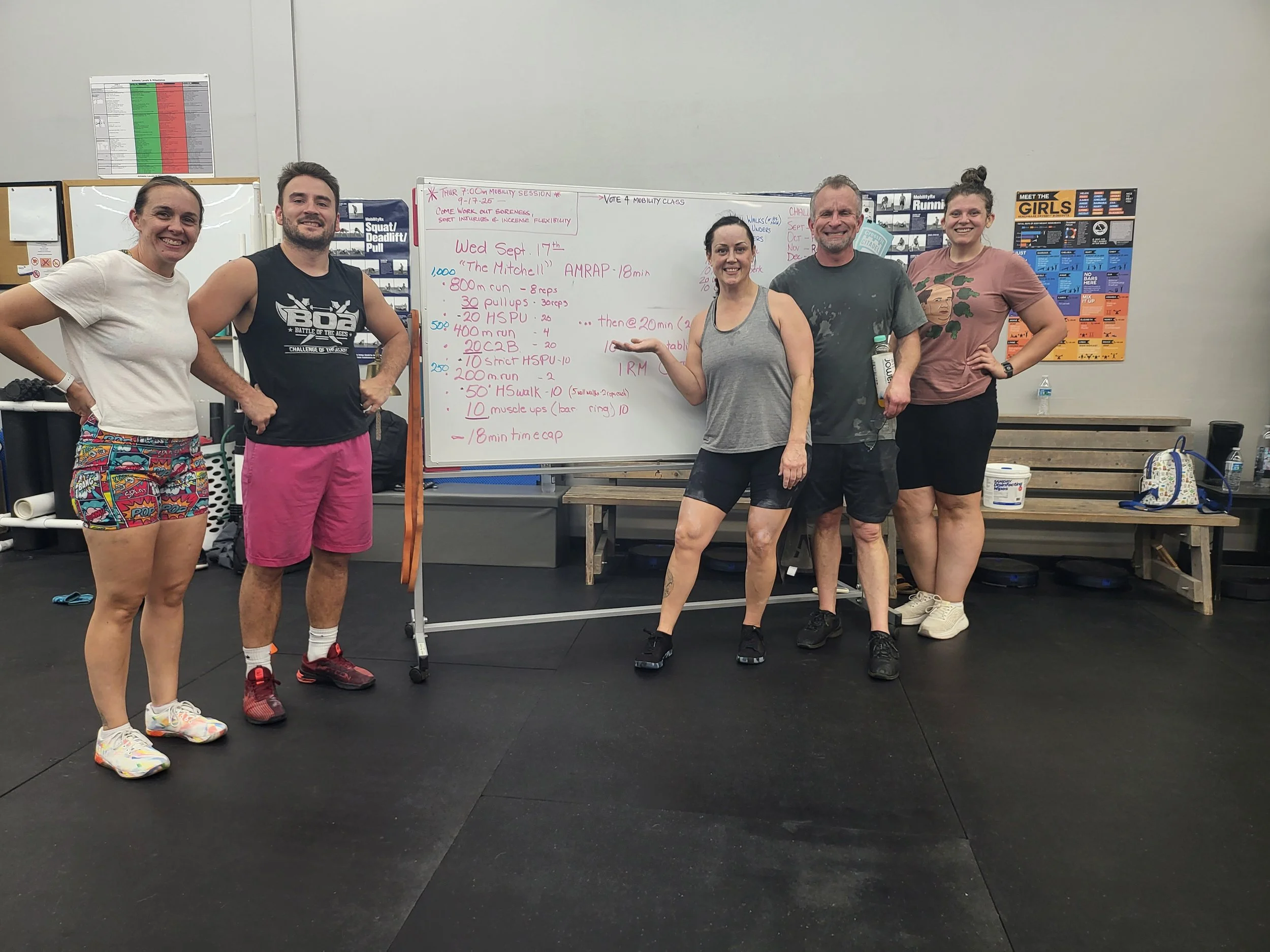 Group of five people standing in a gym in front of a whiteboard with workout notes, smiling and posing for the photo.