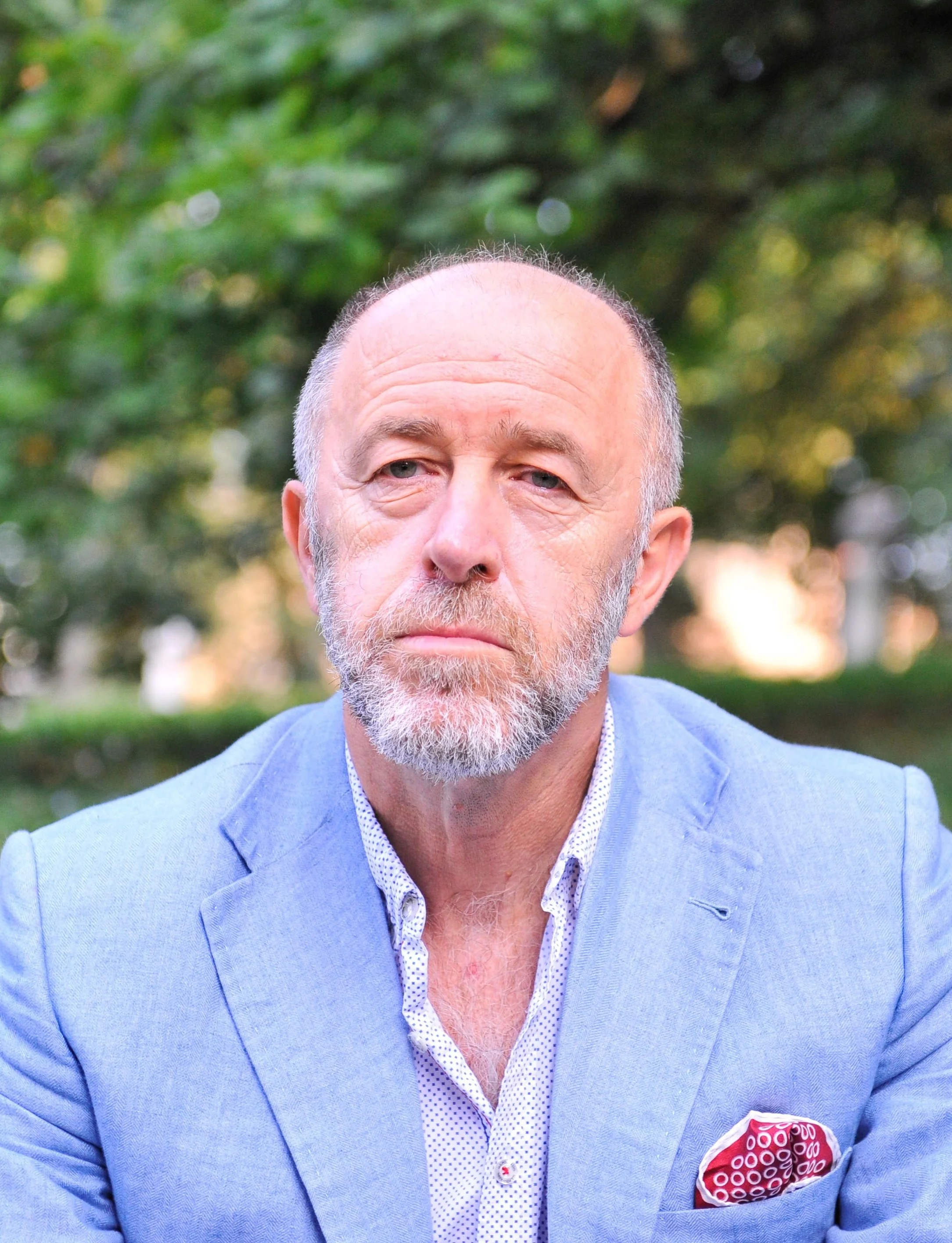 Close-up of a middle-aged man with a bald head and gray beard, wearing a light blue blazer and white shirt with small blue dots, standing outdoors with green trees in the background.