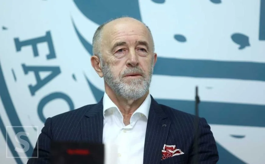 A mature man with a beard and bald head dressed in a dark pinstripe suit and white shirt, sitting in front of a blue and white background with a partially visible logo.