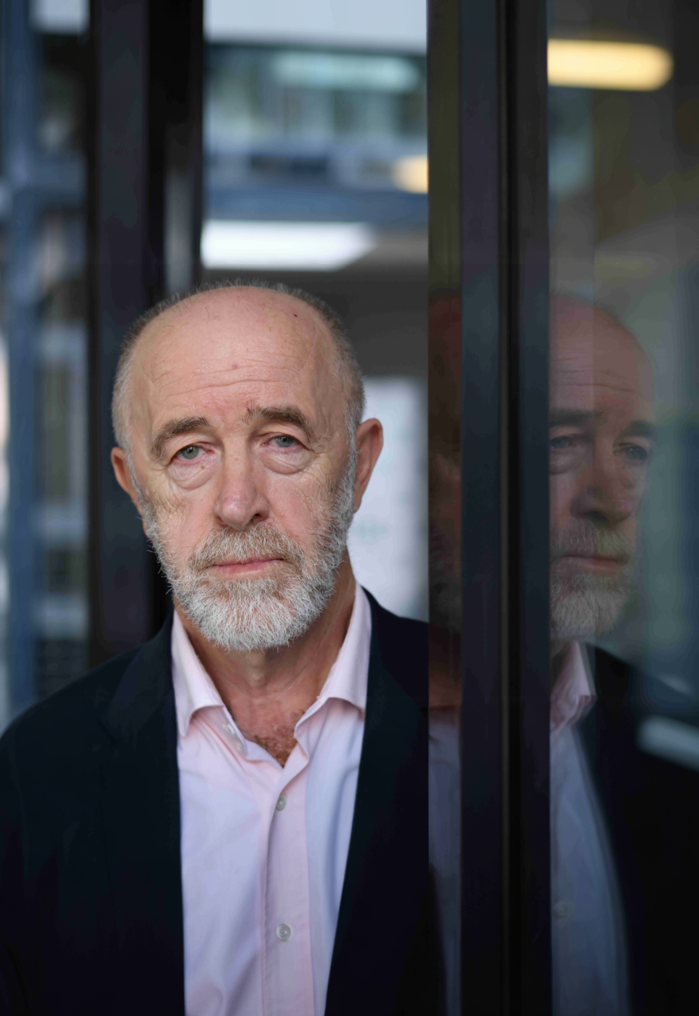 An older man with a white beard and mustache standing behind a glass window, wearing a black jacket and a white shirt, with a serious expression.