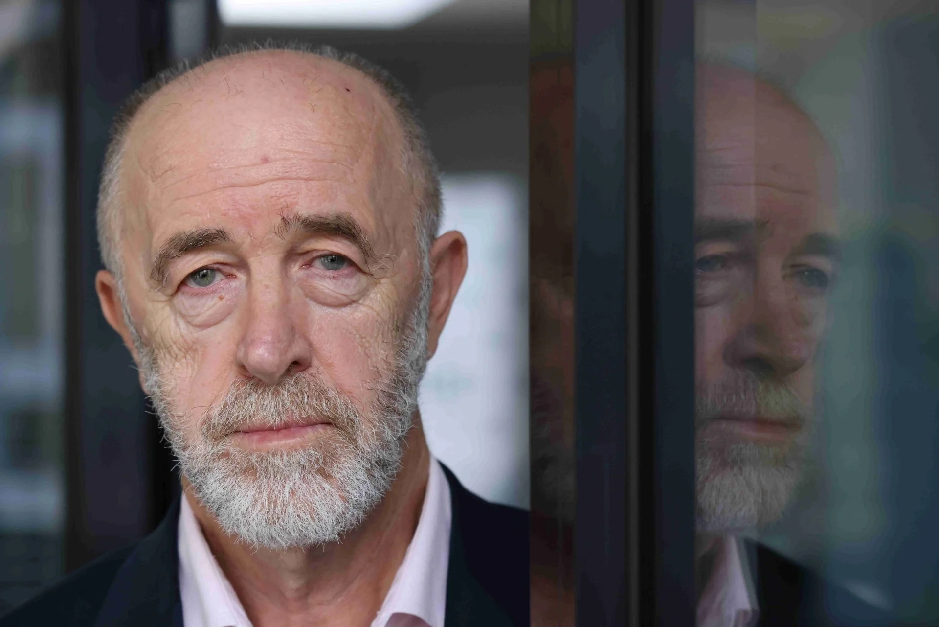 Close-up of an older man with a gray beard and bald head standing next to a glass of reflective surface, showing his reflection.