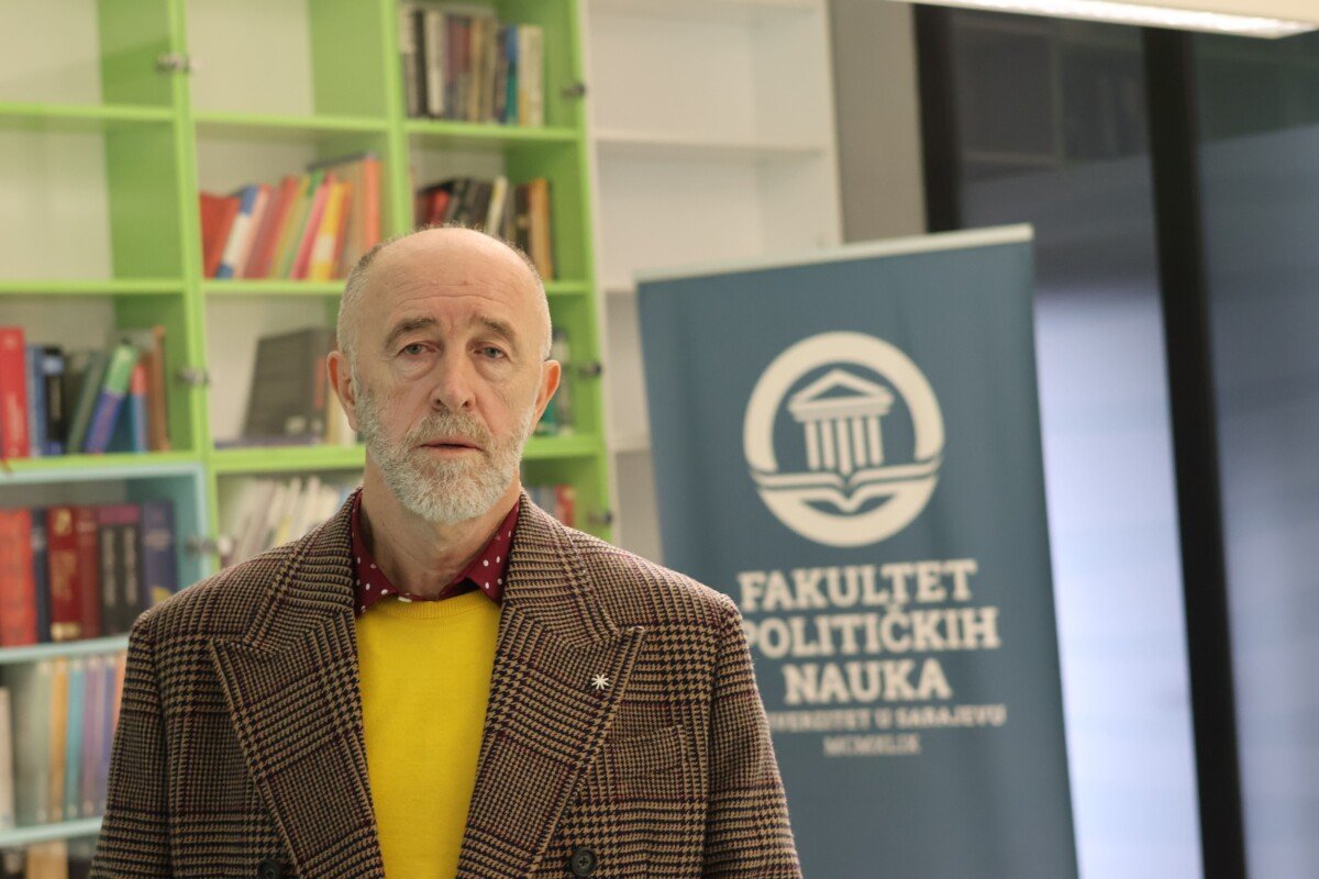 An older man with a gray beard and balding head standing in front of a green bookshelf filled with books, and a blue banner with white text and a logo that reads "FACULTET POLITIČKIH NAUKA".