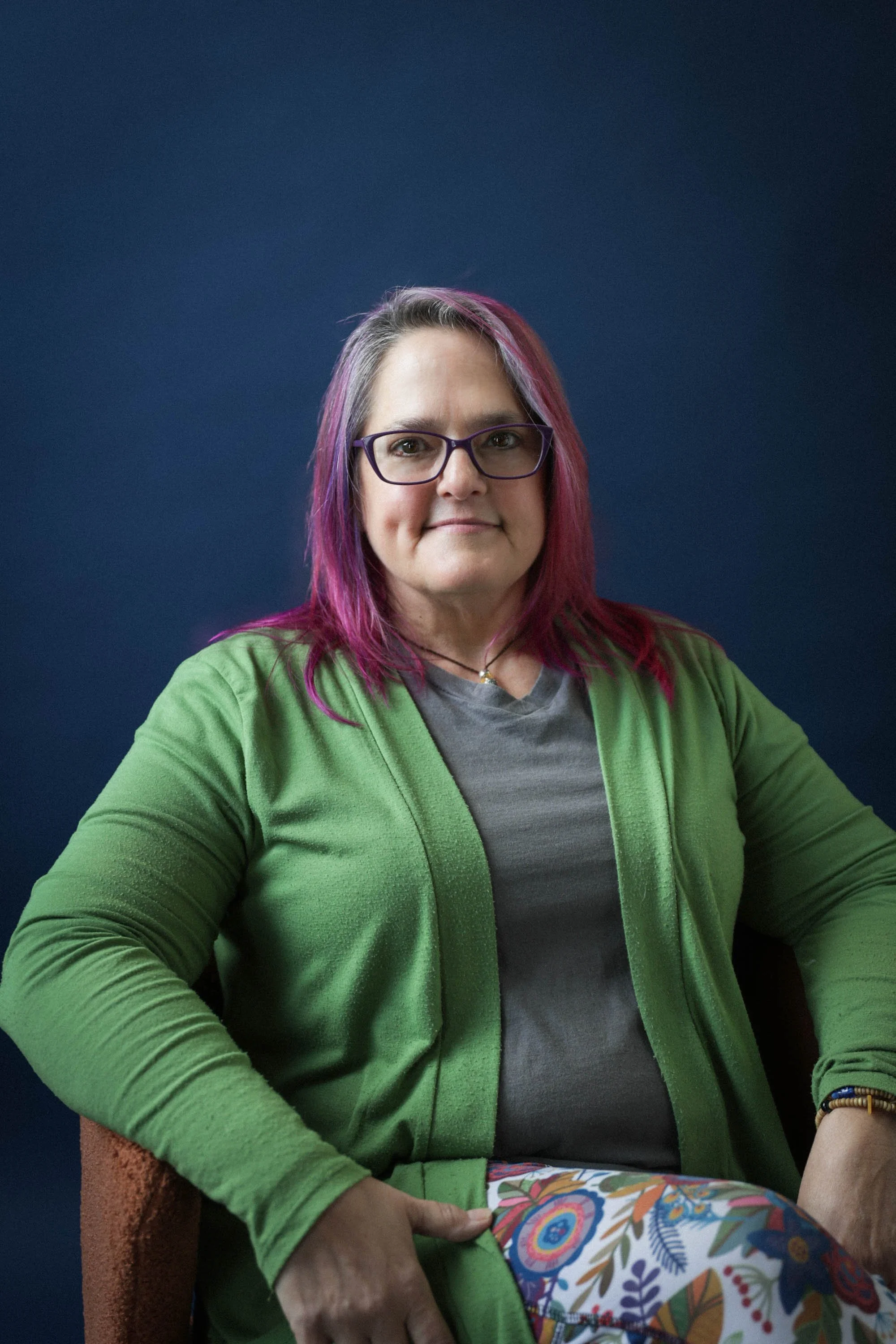 Joyelle Koch - A woman with purple and pink hair wearing glasses, a gray shirt, and a green cardigan, sitting on a chair against a dark blue background.