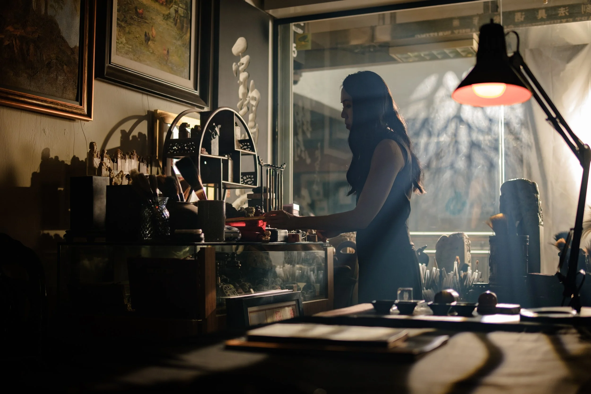 A woman with long dark hair behind a desk gathering art materials, with paintings hanging on the wall behind her, warm sunlight streaming through a large window, and art supplies on the desk.