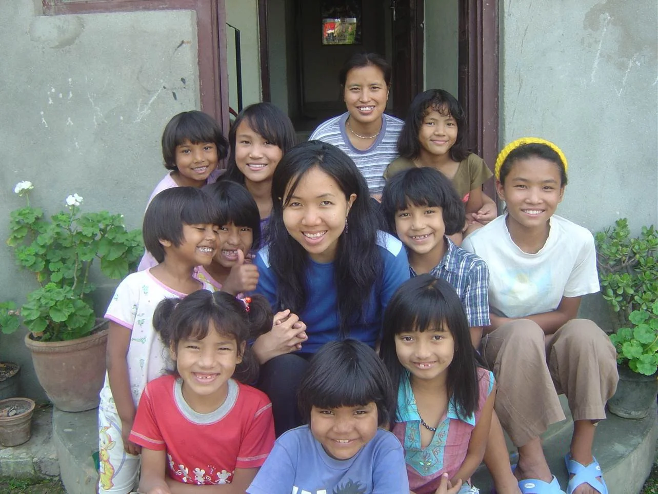 Group of smiling children and two adult women posing outside in front of a concrete building with potted plants nearby.