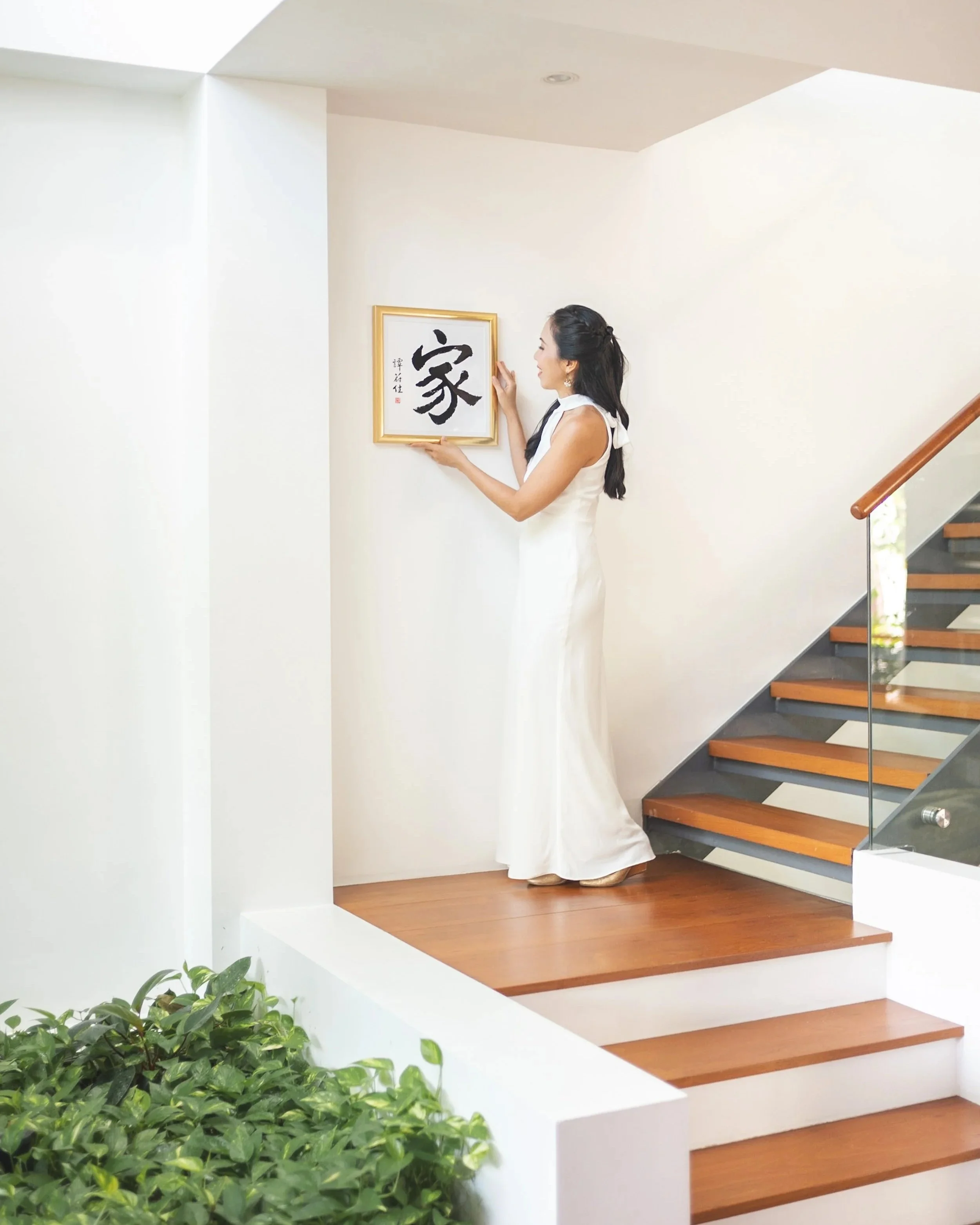 Woman in white dress standing near a wall, touching a framed artwork with a black Chinese character, staircase on the right, green plants in the foreground.