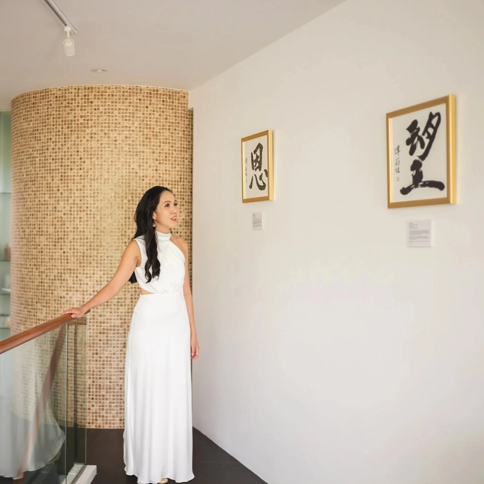 Woman in a white dress standing in an art gallery, looking at framed calligraphy on the wall.
