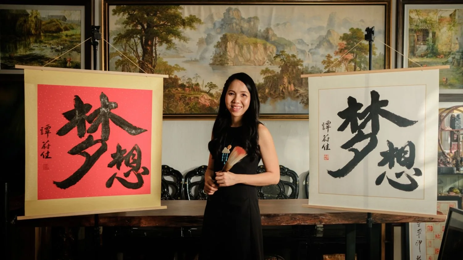 Wai-Jia Tam standing between two calligraphy scrolls with Chinese characters, holding a fan and brush, in front of a landscape painting in an art gallery or studio.