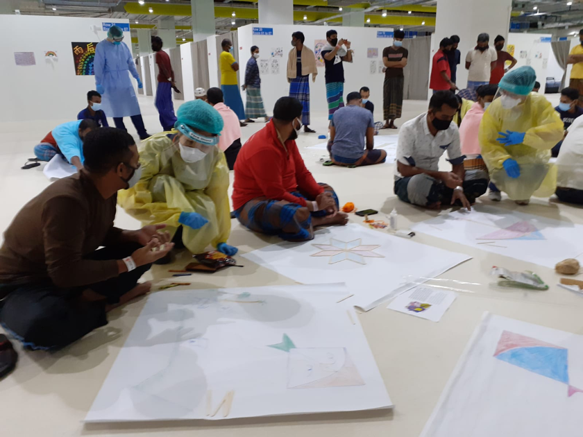 People sitting on the floor creating colorful geometric artwork at an indoor event, some wearing protective gear, with others standing and observing in the background.