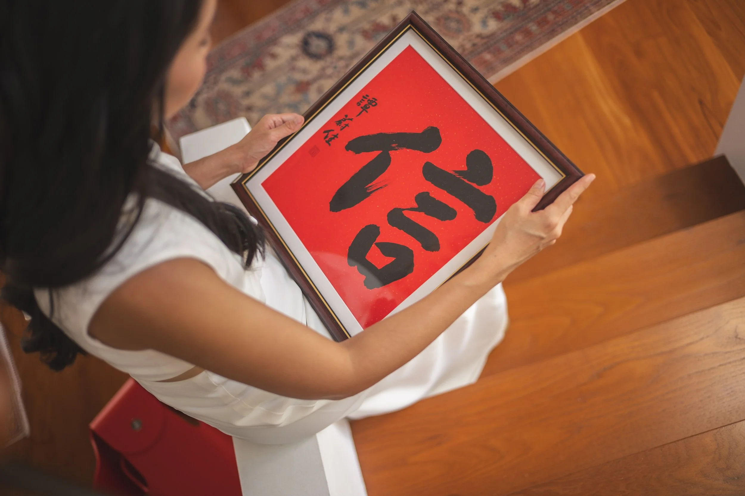 A woman in a white dress holds a framed artwork with a red background and black calligraphy.