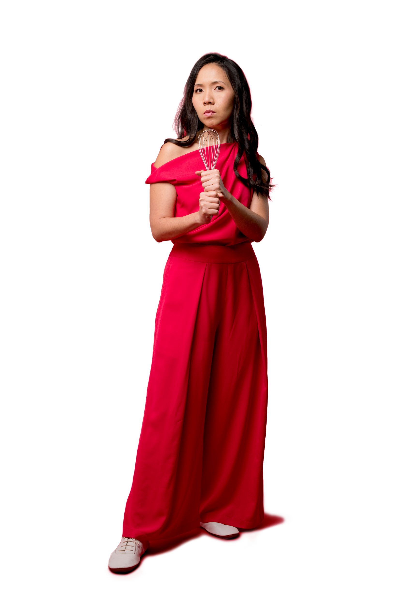 Wai-Jia Tam in a red jumpsuit and white shoes stands against a red background, holding a metal whisk in front of her.