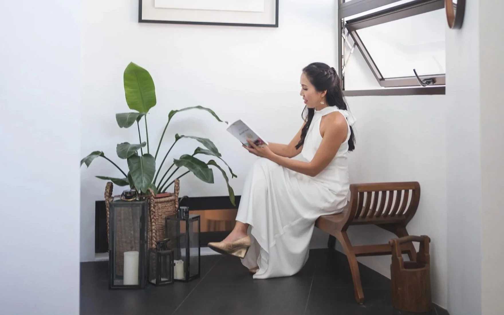 Woman in a white dress reading a book while sitting on a wooden bench in a minimalist room with a large green plant and decorative lanterns.
