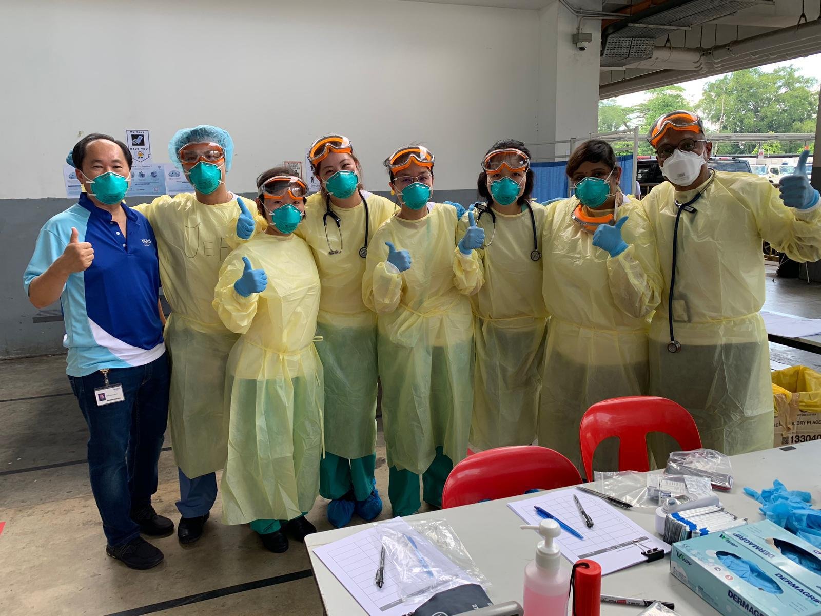 A group of healthcare workers and a visitor wearing protective masks, gloves, and gowns, giving thumbs-up gestures inside a medical facility.