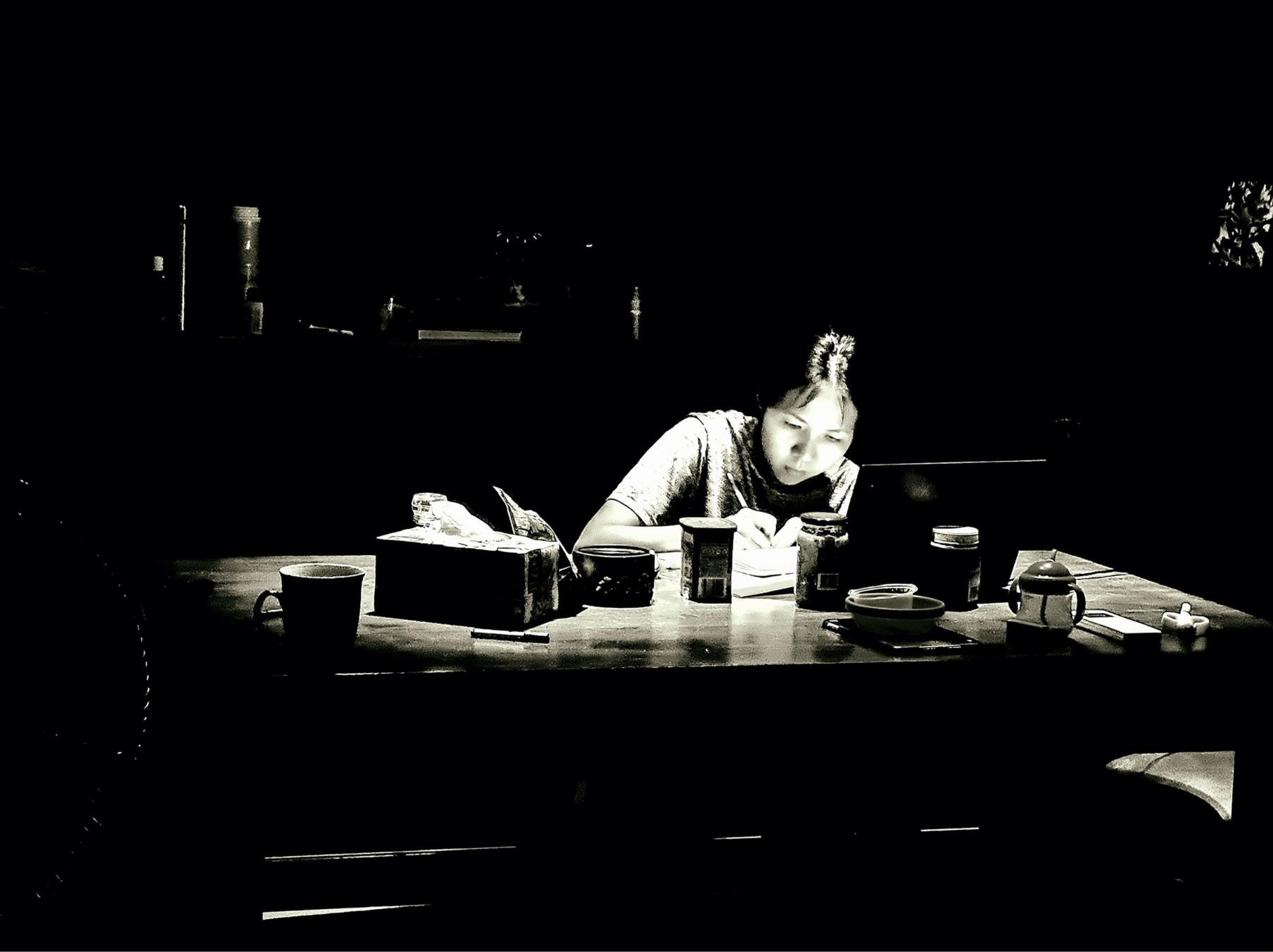A woman sitting at a table, drawing, with various objects such as cups, containers, and a laptop on the table, illuminated by a light source.