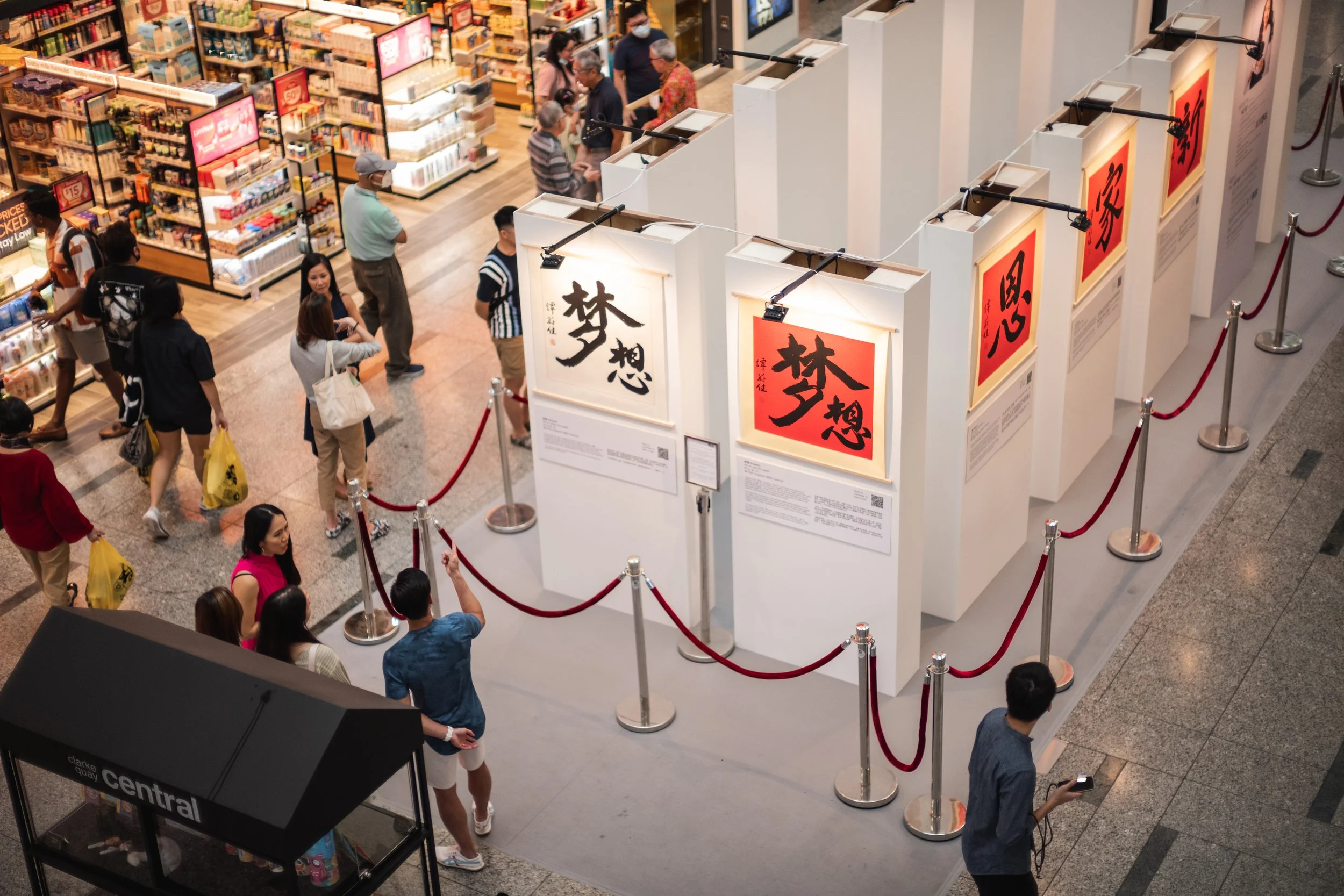 Several people are shopping and browsing in a bookstore or section of a retail store, with display boards featuring Chinese calligraphy scrolls as part of an art exhibit.