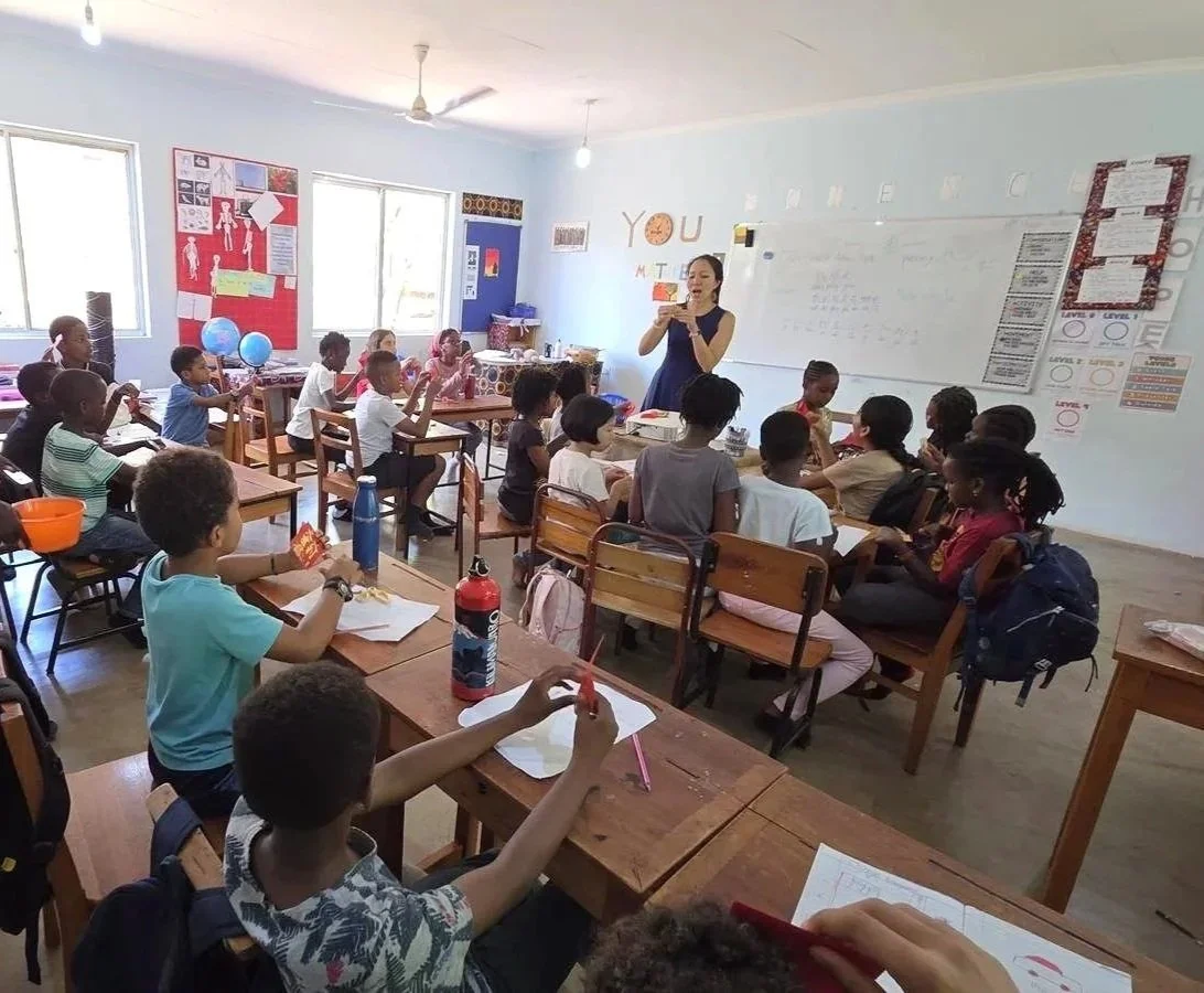A classroom with a teacher standing at the whiteboard and teaching students sitting at desks, with educational posters on the walls.