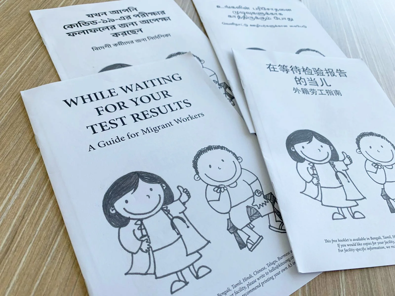 Pamphlets titled 'While waiting for your test results' and some in different languages, featuring cartoon illustrations of children in school uniforms holding thumbs up, placed on a wooden surface.