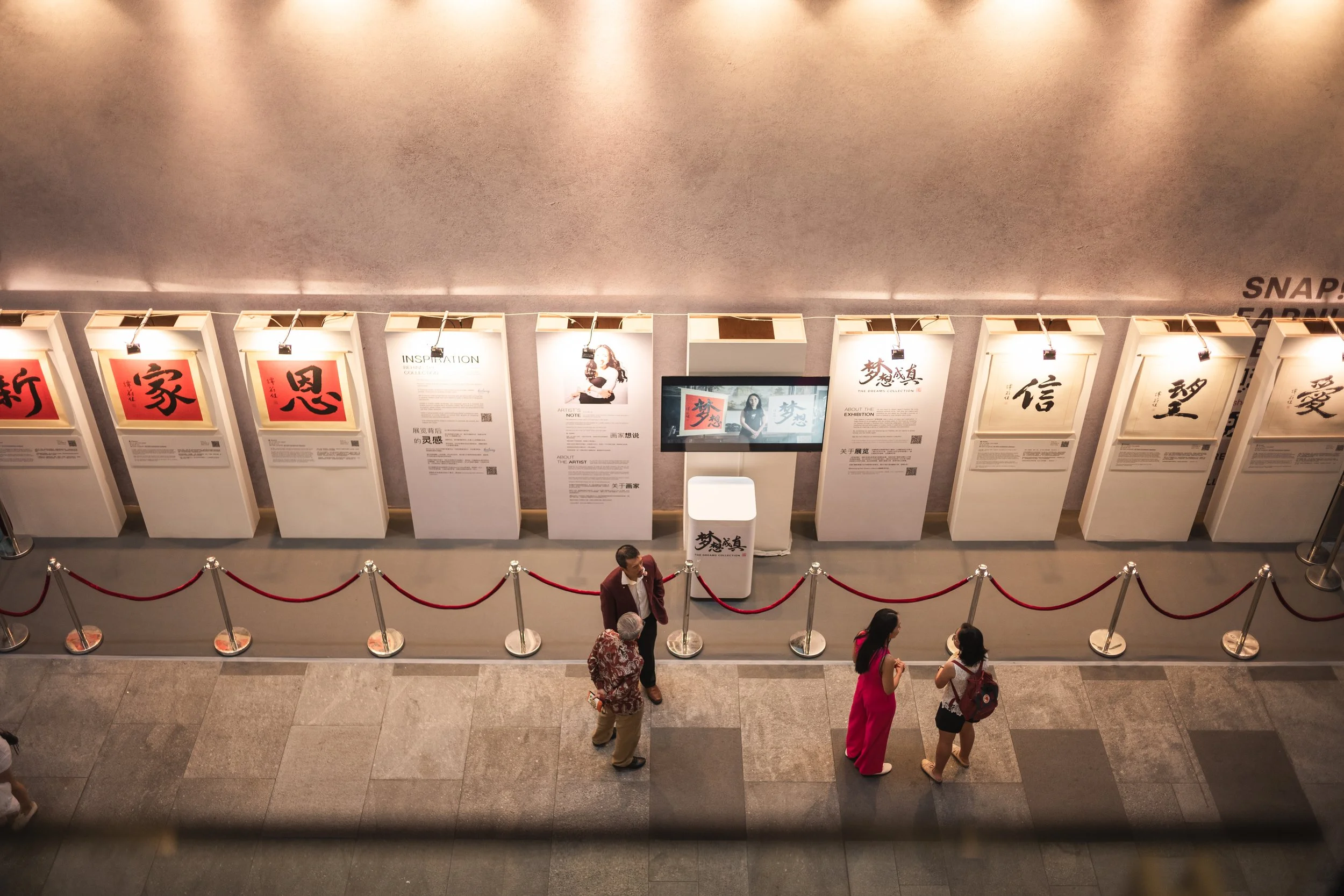 An indoor exhibit with posters featuring Chinese calligraphy and a central screen showing similar artwork. Visitors are walking along a roped off area, observing the displays.