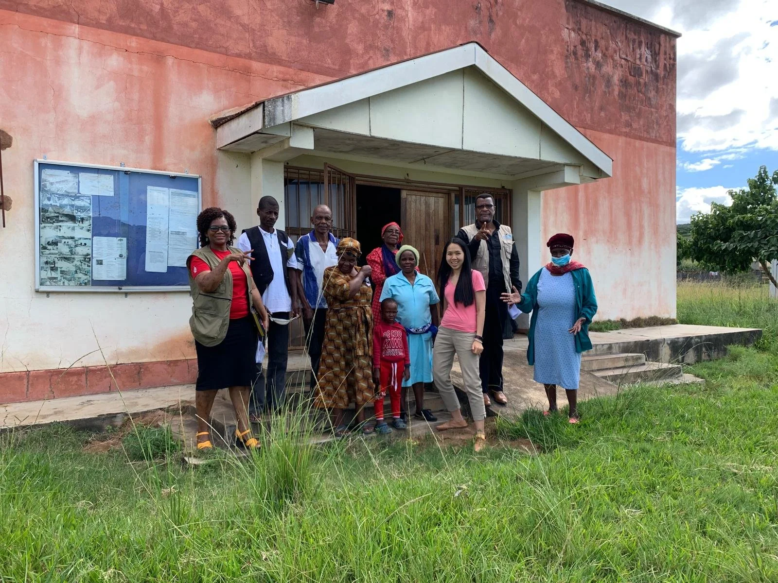 Group of diverse people standing outside in front of a building, posing for a photo. Some are smiling and making gestures.