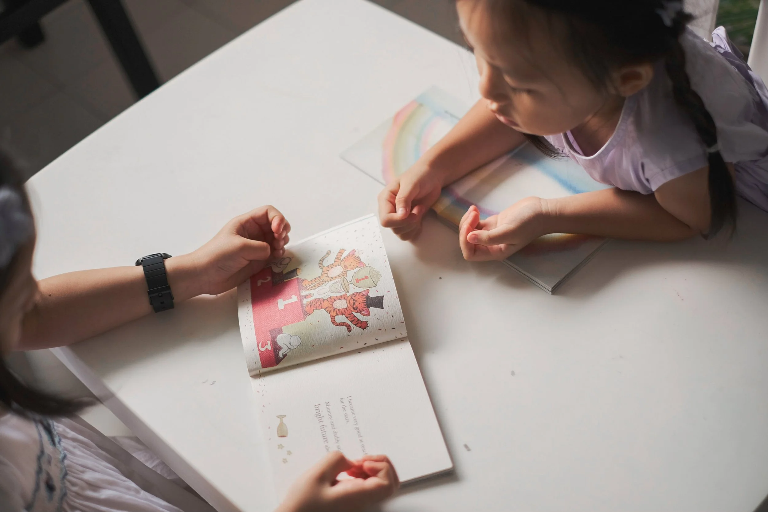 A young girl with dark hair and a braid, wearing a light purple shirt, is looking down at a book or card with colorful illustrations of a tiger and other animals on a white table. An adult's hand, wearing a black wristwatch, is helping her hold the item.