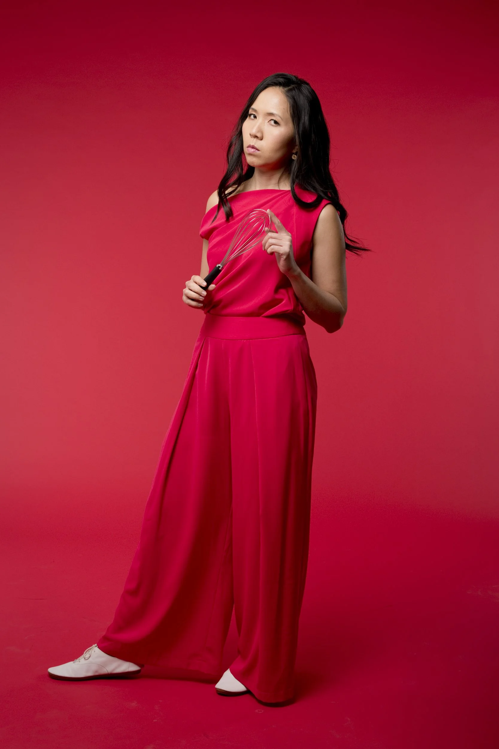 Woman in red dress holding a whisk, standing against a red background, wearing white shoes.