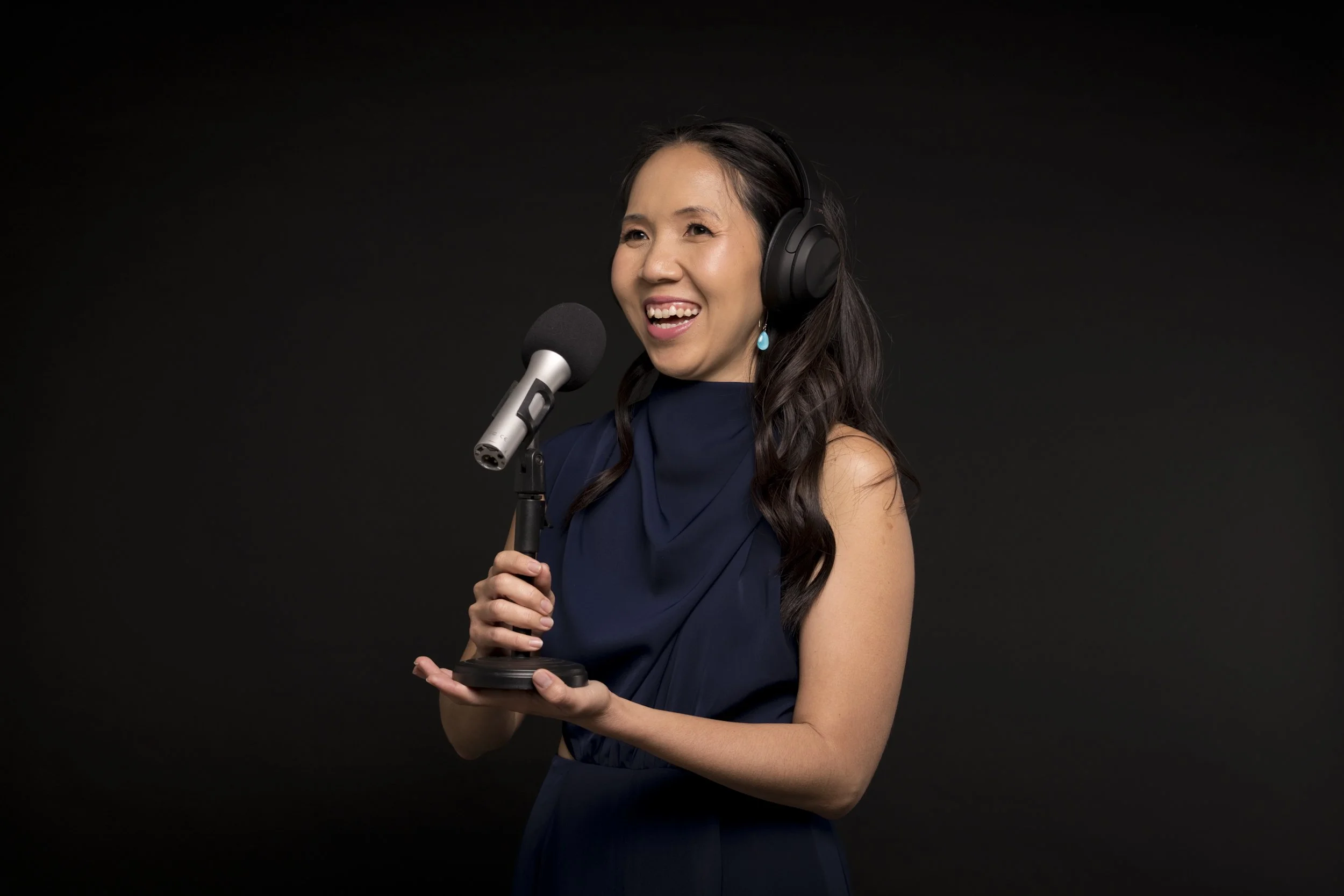 A woman in a navy blue dress is smiling and holding a microphone with both hands against a black background.