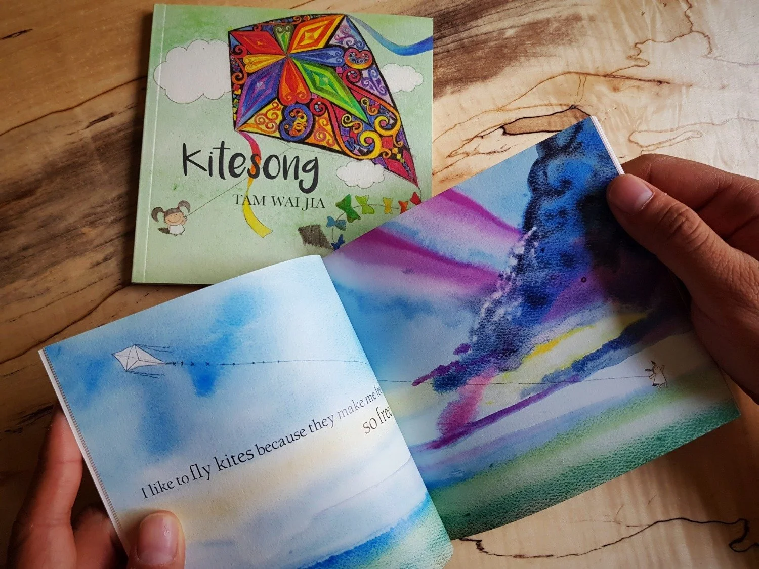 A children's book titled 'Kitesong' by Tam Wai Jia is open, displaying colorful illustrations of a kite flying in the sky, with a girl holding the kite string. The right page features a watercolor painting of a kite in the sky and a paragraph of text about flying kites.