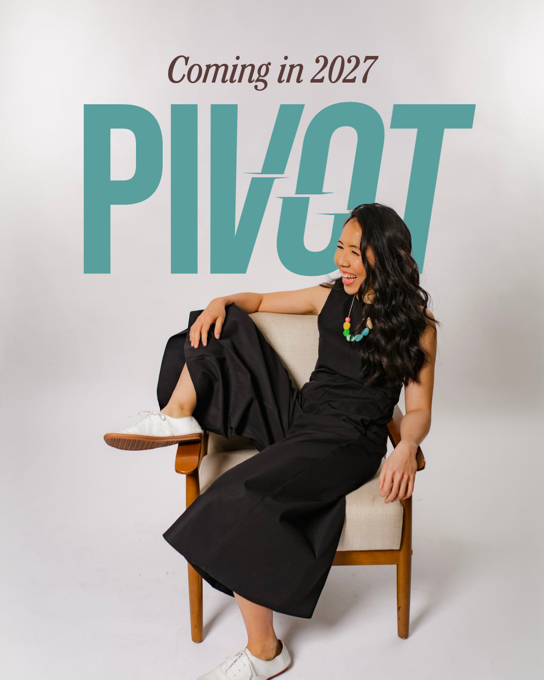 A woman with long, curly black hair sitting on a chair with one leg crossed over the other, laughing. She is wearing a black sleeveless dress, white sneakers, and a colorful necklace. Behind her is a wall with a large text graphic that says 'Coming in 2027 PIVOT'.