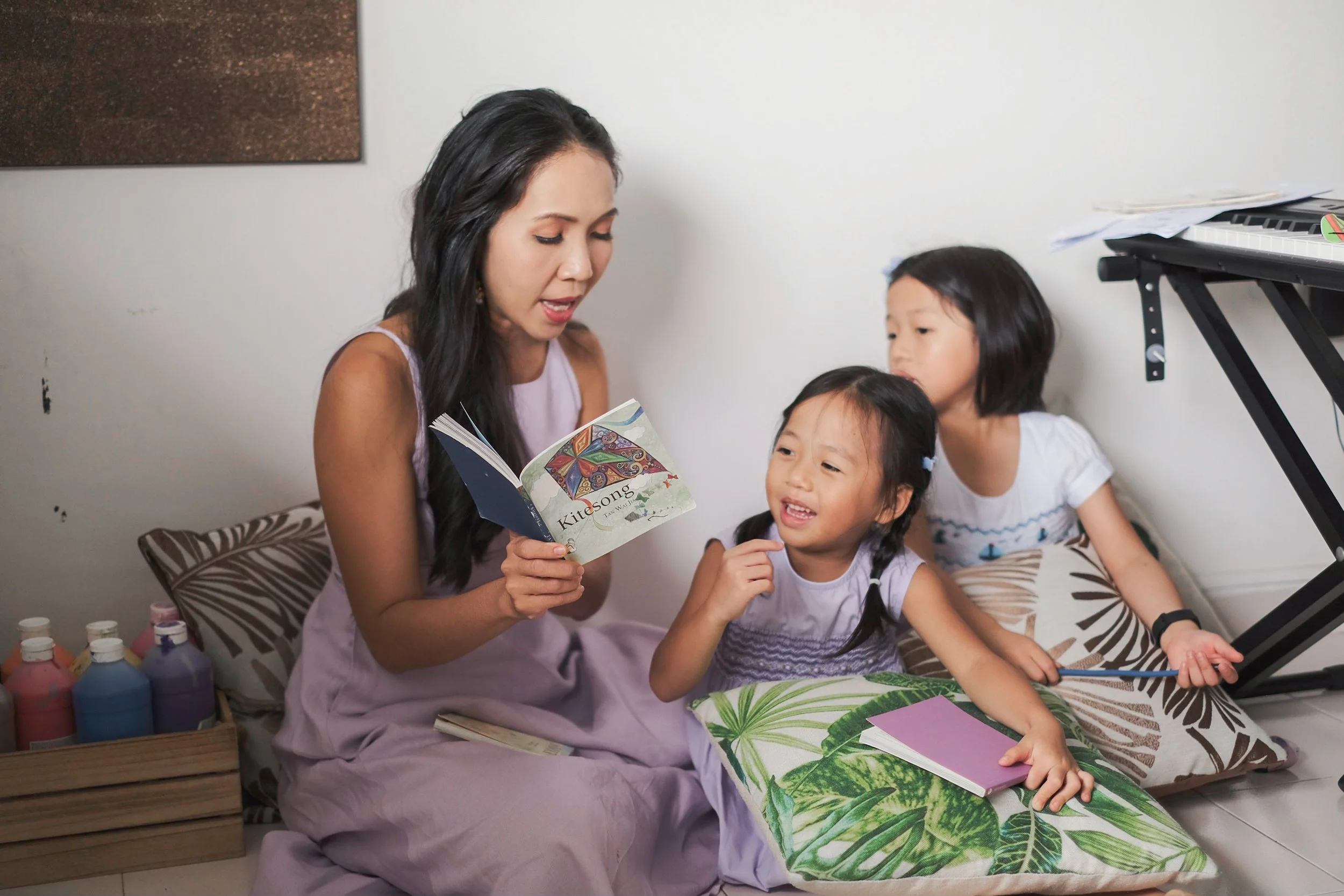 A woman reading a book to two young girls sitting on the floor in a cozy room with pillows, books, and art supplies, near a keyboard on a stand.