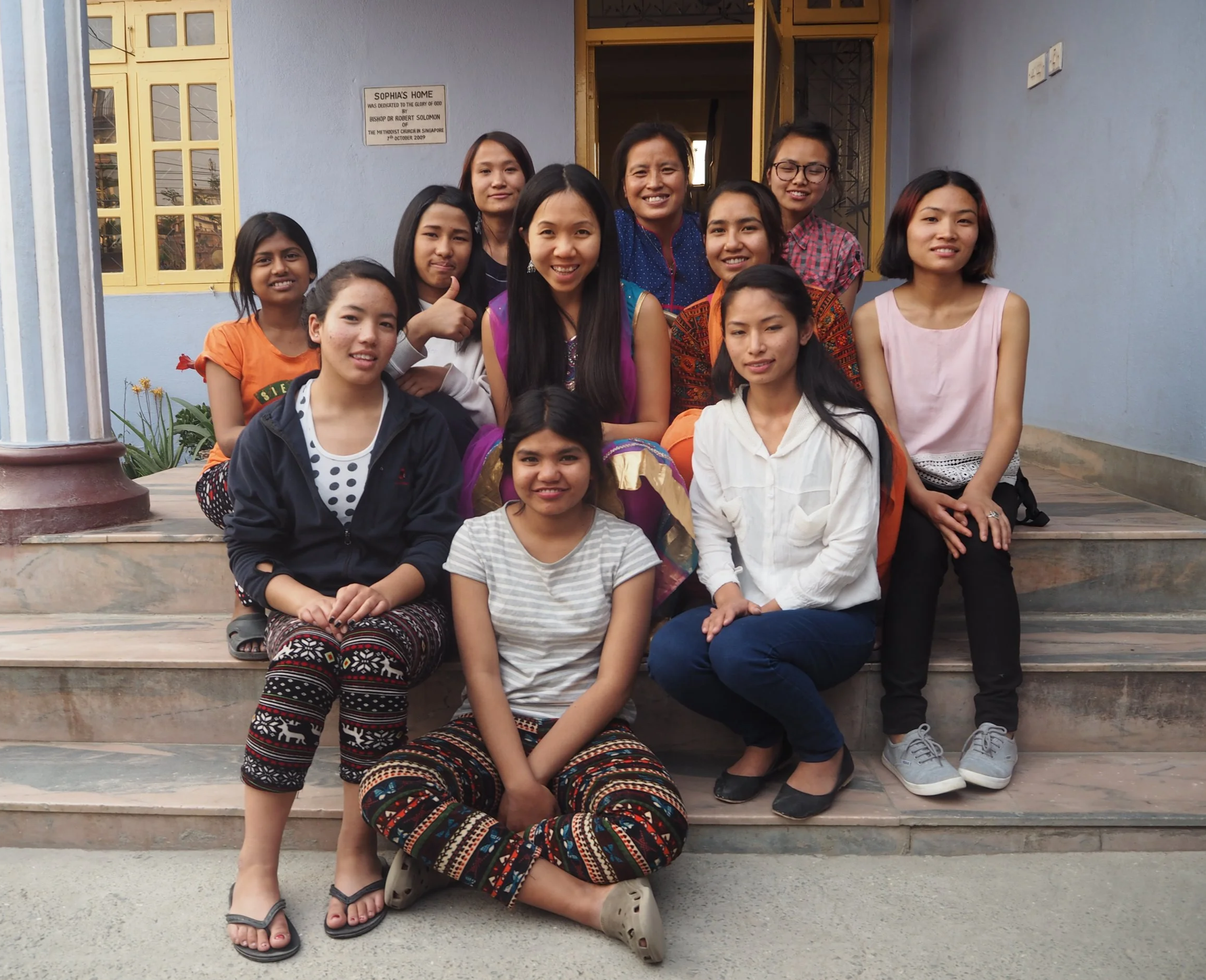 A group of women and girls standing and sitting on stairs outside a building with a blue wall and yellow window frames, smiling for the photo.