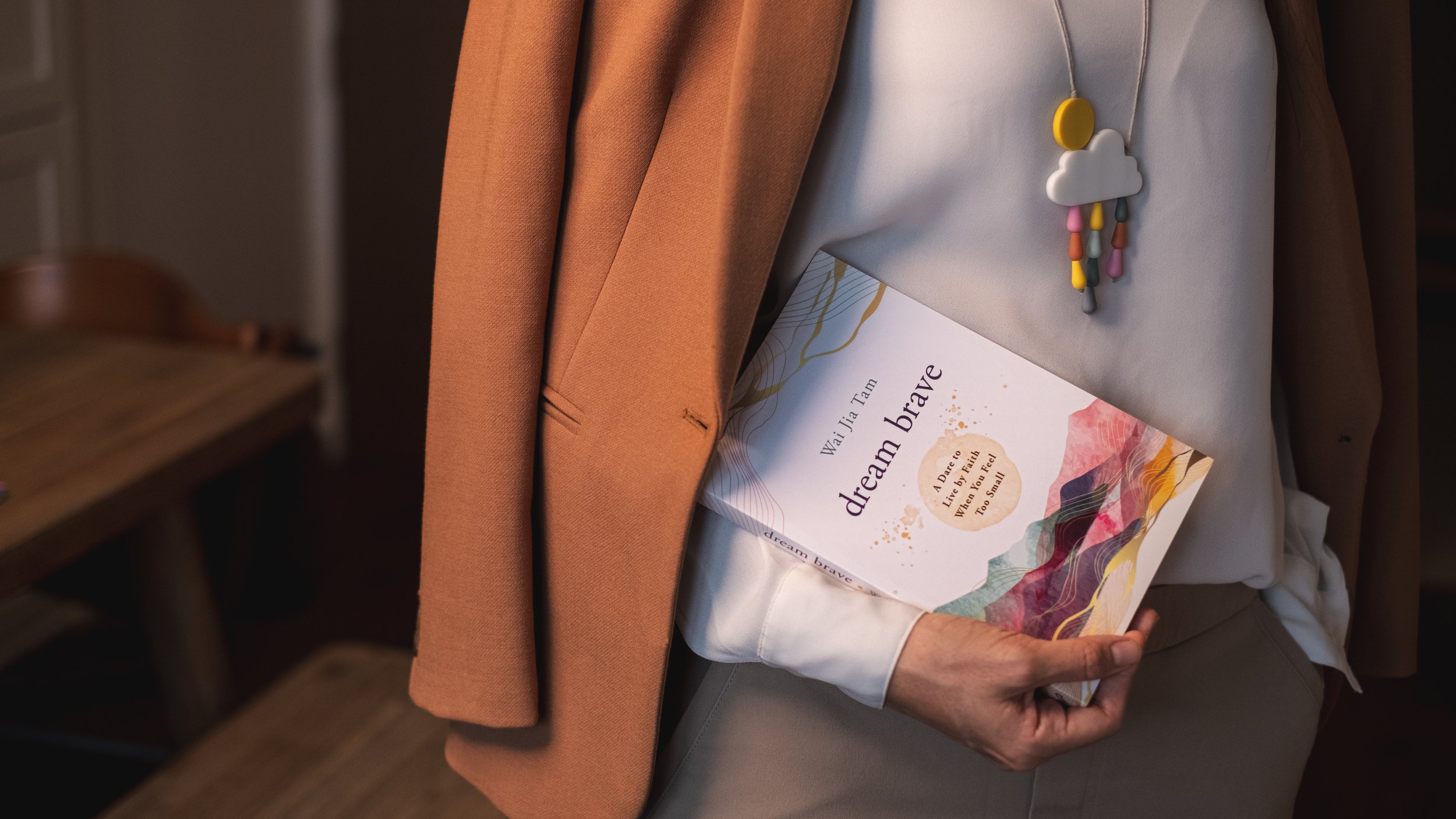 Person holding a book titled 'Dream Brave' by Wai-Jia Tam, wearing a white top and a necklace with a cloud charm, with a tan blazer draped over their arm.