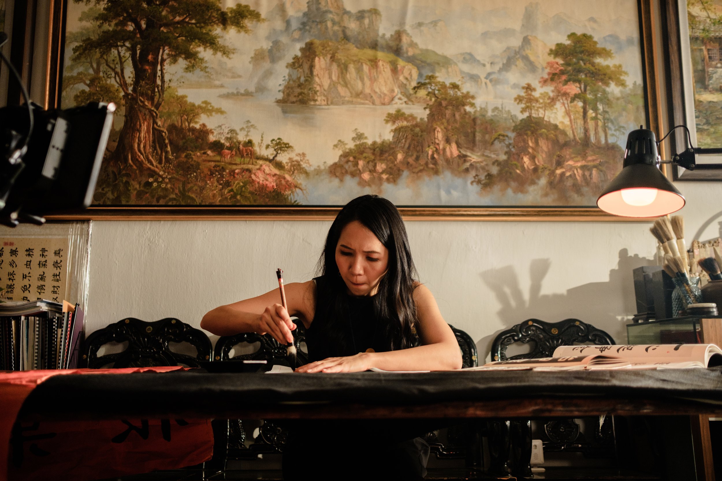 A woman with long black hair sitting at a table, writing or drawing with a brush and ink in a restaurant or cafe. Behind her, a large landscape painting of mountains, trees, and a body of water adorns the wall. There are lamps and various items on th