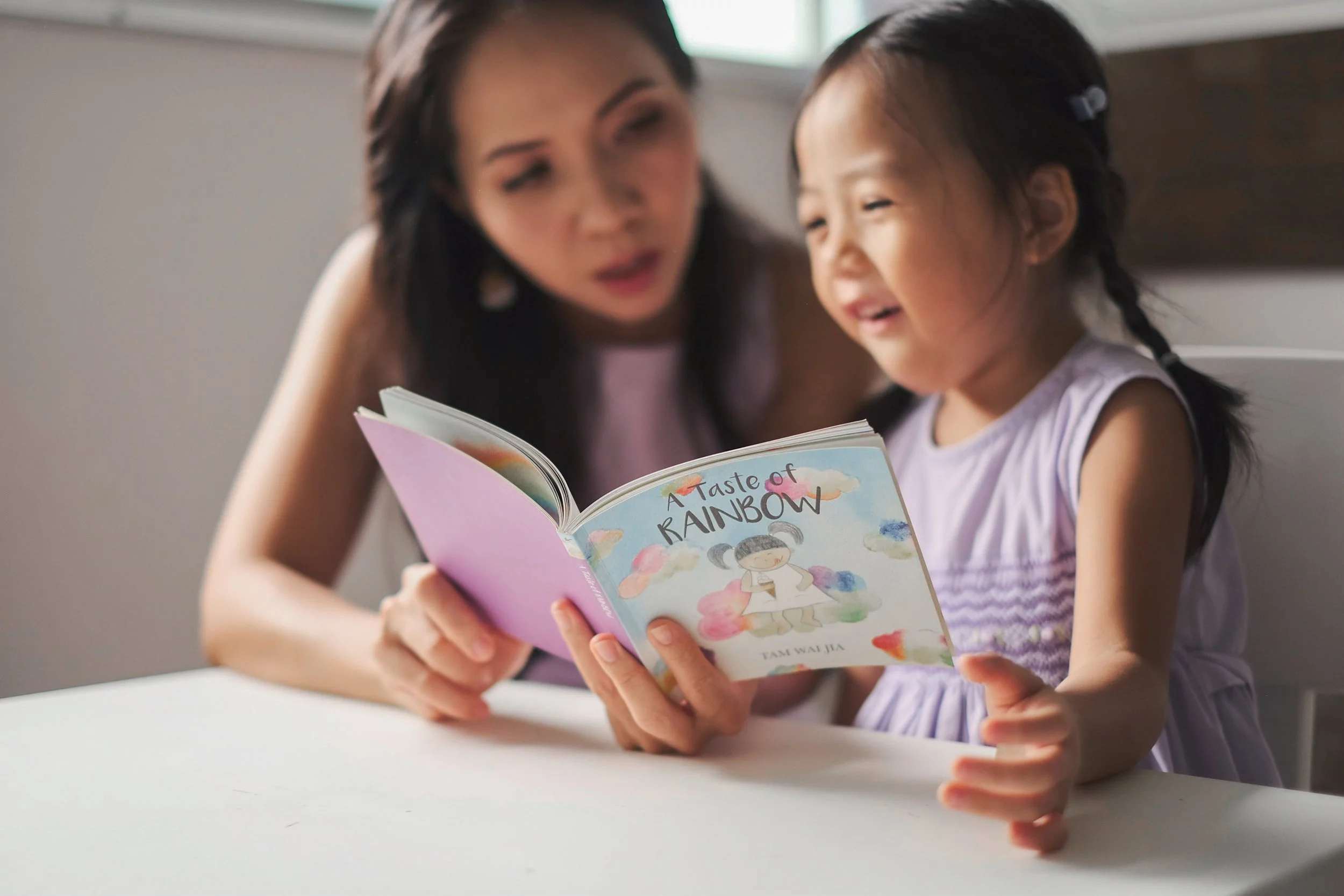 A woman and a young girl reading a colorful children's book titled 'A Taste of Rainbow' together at a table.