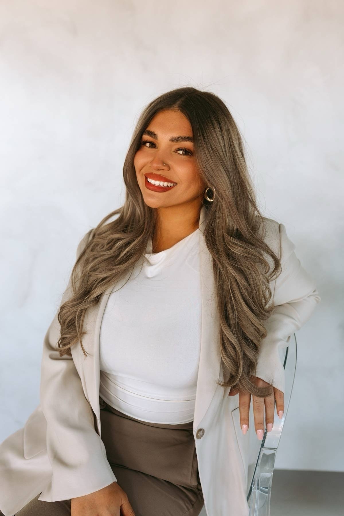 A woman with long wavy hair, wearing a white blazer and a white top, sitting on a transparent chair, smiling at the camera against a plain, light-colored background.