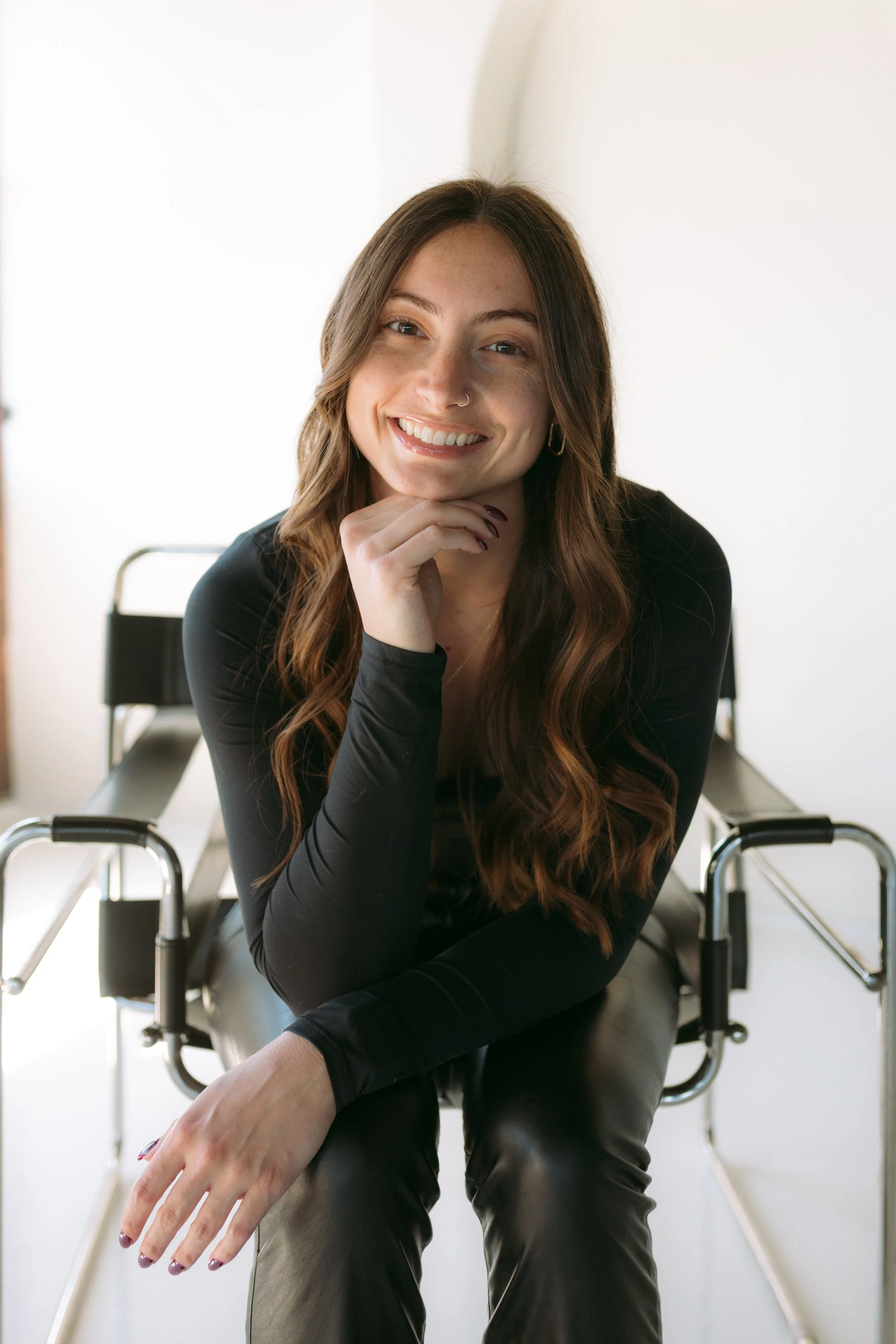 A young woman with long, wavy brown hair, wearing a black top and leather pants, sitting on a wheelchair, smiling and looking at the camera with one hand resting on her chin.
