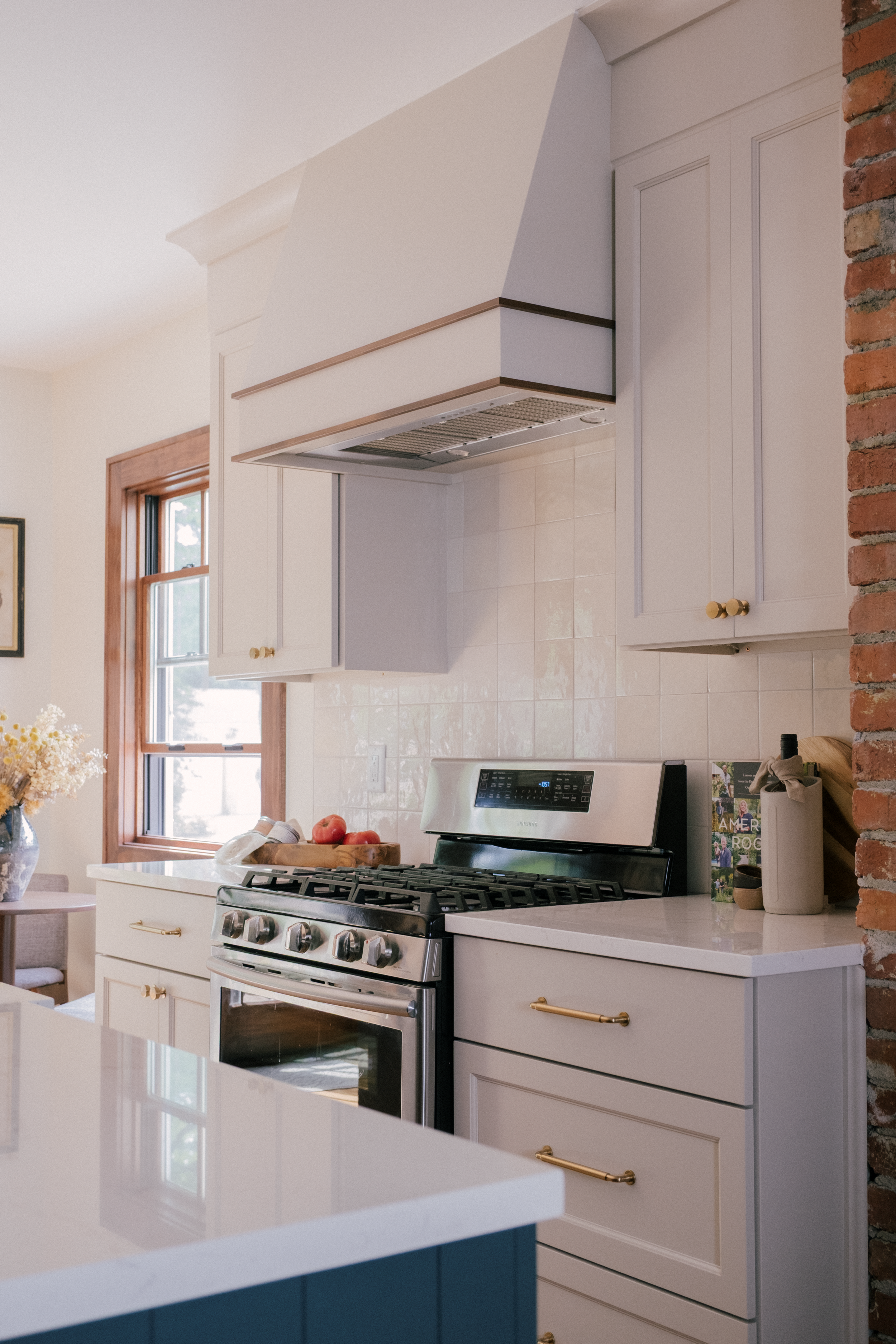 A cozy kitchen with white cabinets, a white range hood with decorative accents, and a brick accent wall. Sunlight streams in through a window, creating a warm, inviting atmosphere.