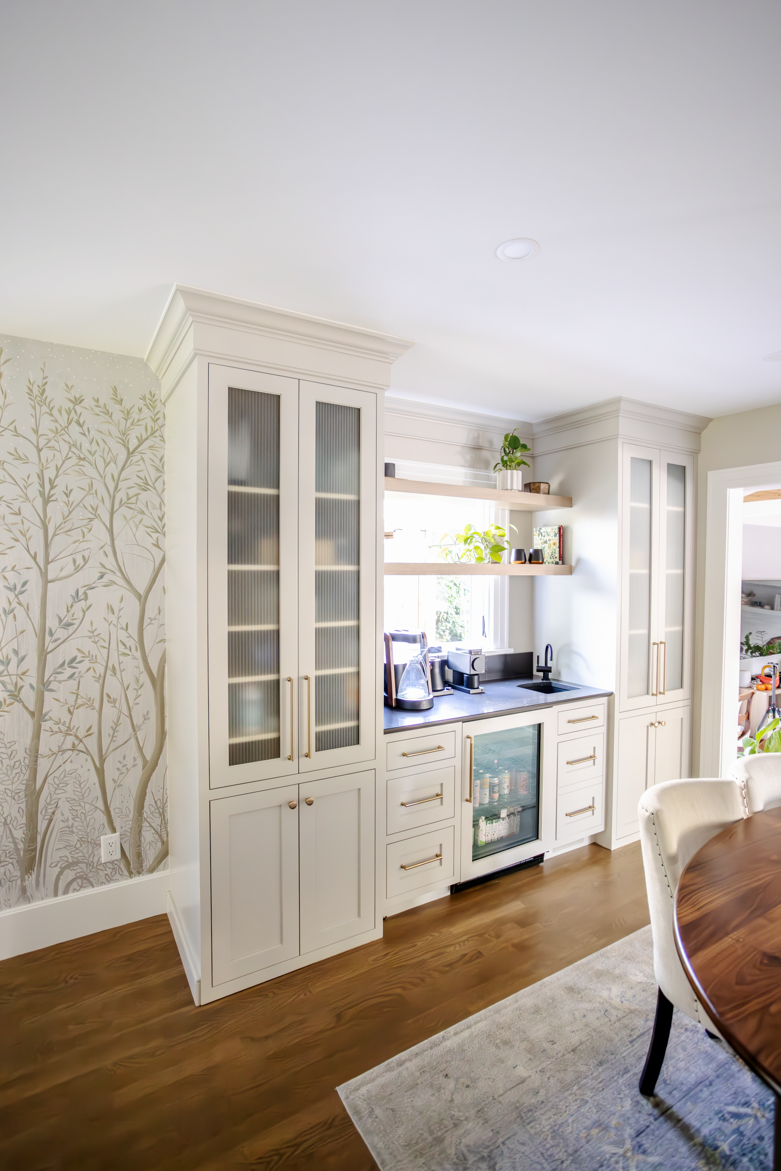 Bright wet bar with white cabinetry featuring reeded glass panels, natural wood open shelving, and a beverage fridge. Shelves hold plants, adding warmth. A wall mural showcasing an elegant forest scene is visible in the background.