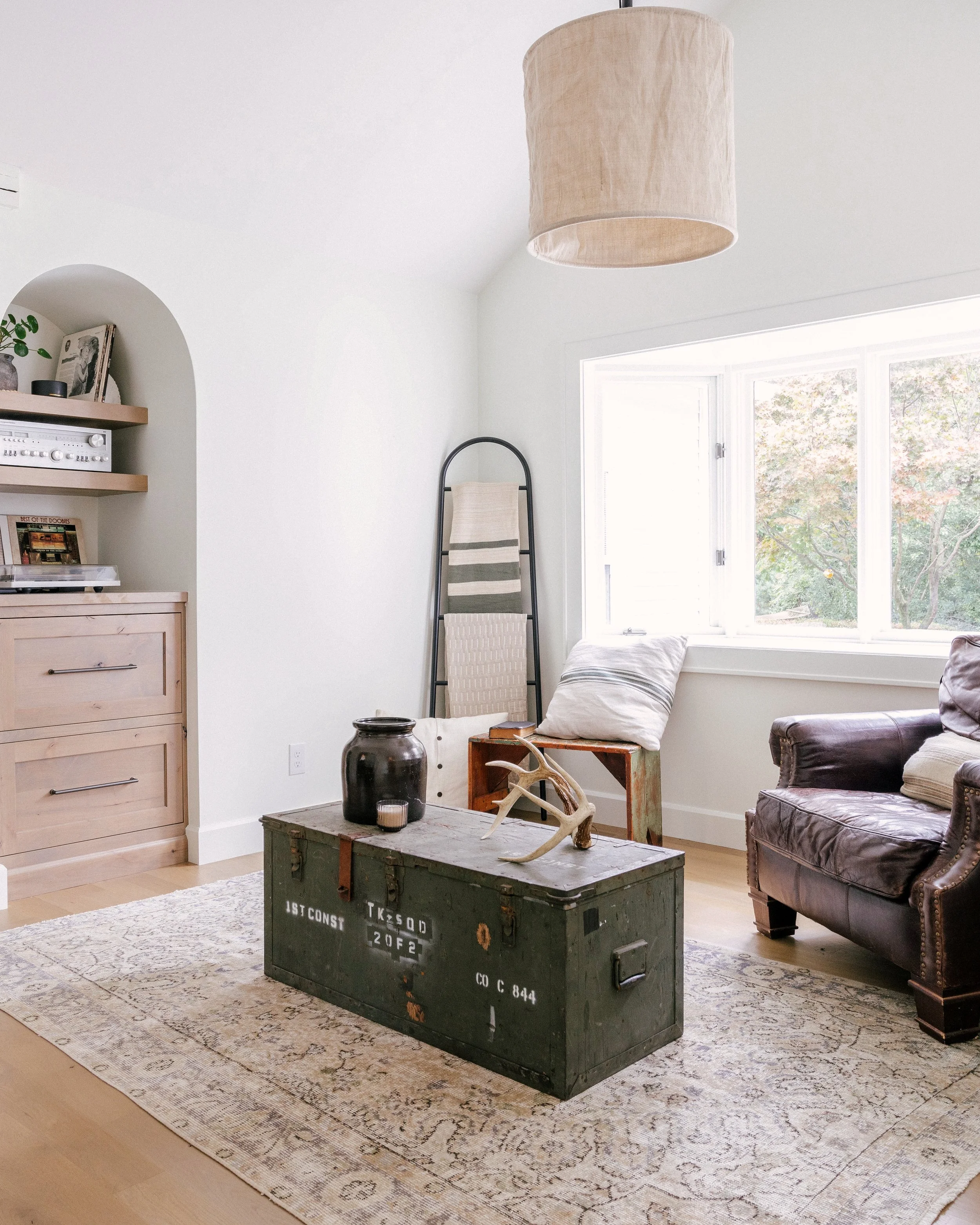 Cozy living room with a vintage trunk as a coffee table, topped with decor. A leather armchair, record player niche, and warm, neutral tones create a welcoming atmosphere.