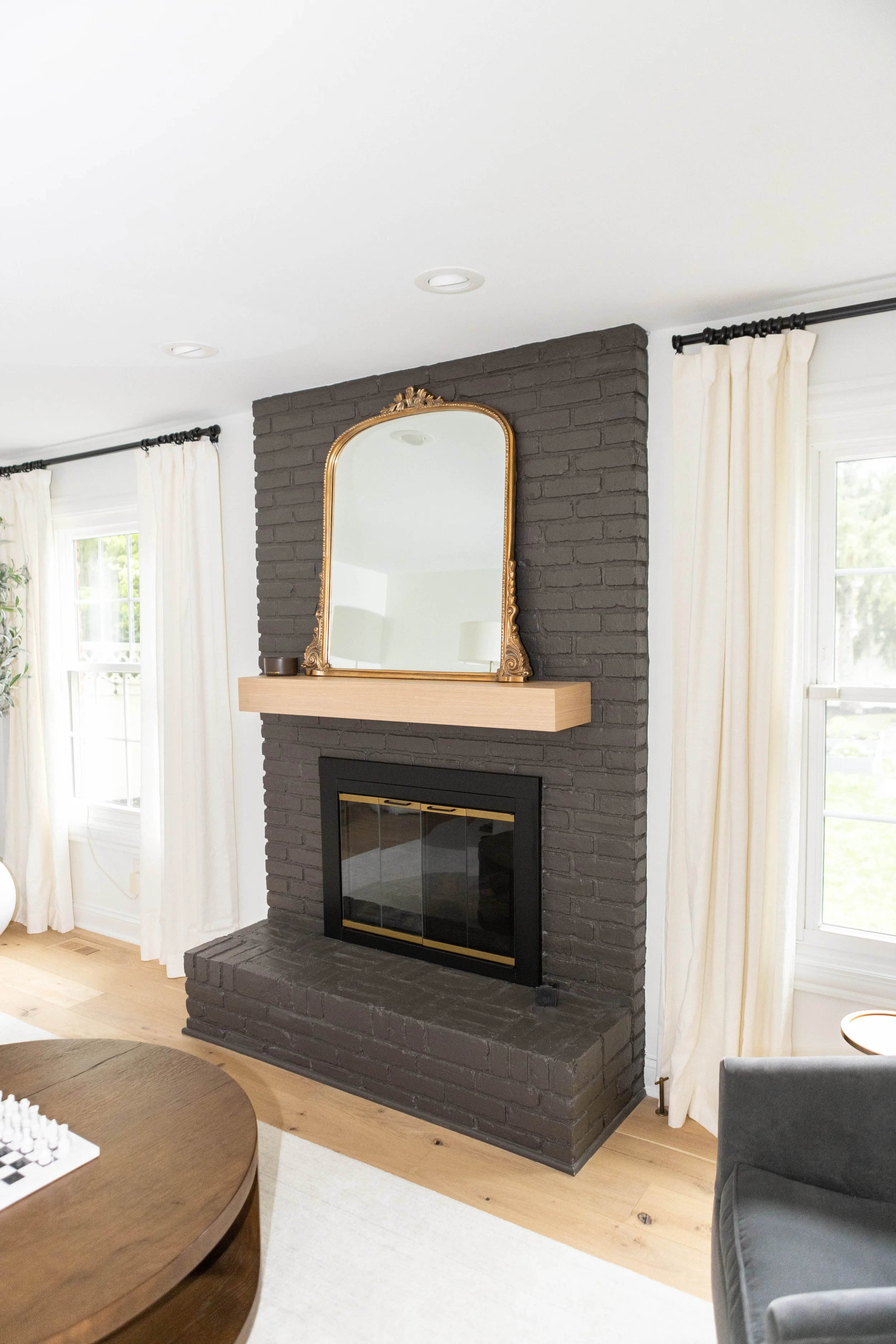 A cozy living room with a painted brick fireplace, wooden mantel, and ornate mirror above. White curtains and light hardwood floors create a bright, elegant atmosphere.