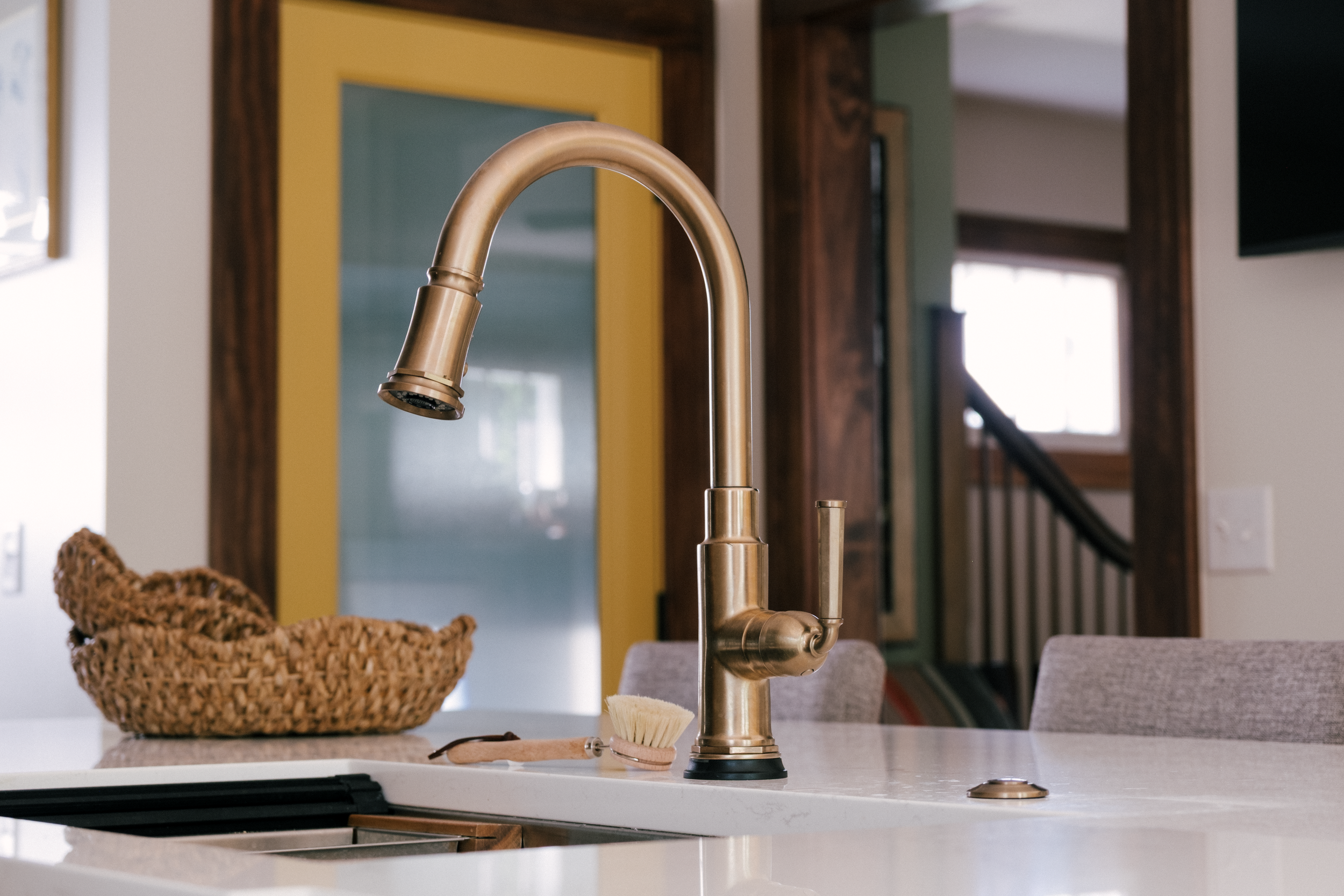 A modern Brizo Rook kitchen faucet on a quartz countertop, with a woven basket and brush nearby. A painted yellow door with inset reeded glass is visible in the background.